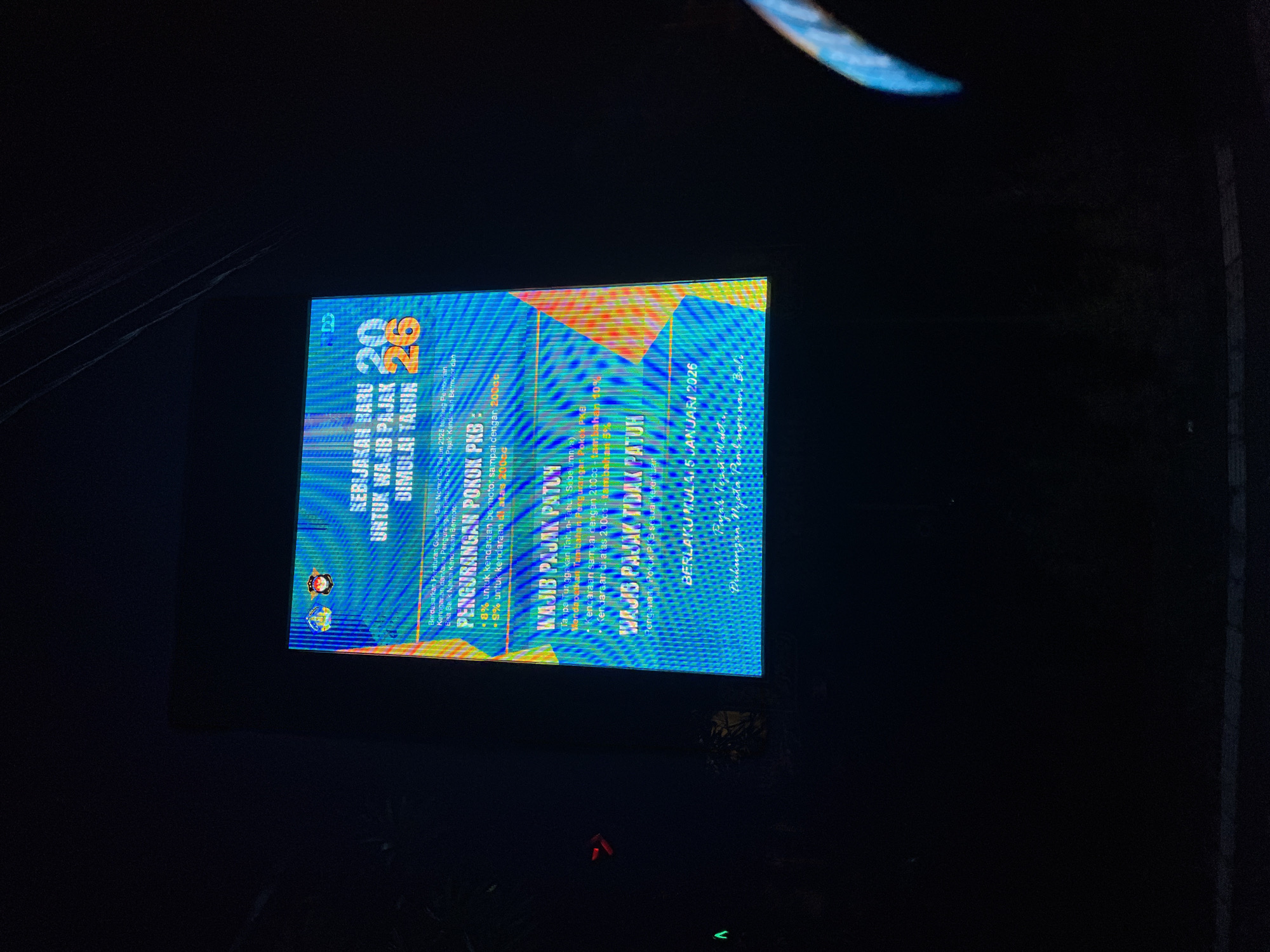 The image shows a digital billboard at night in Denpasar, Indonesia, displaying information about new tax policies. The billboard has a blue and green gradient background with orange accents. The text on the billboard is in Indonesian and reads "KEBIJAKAN BARU UNTUK WAJIB PAJAK DIMULAI TAHUN 2026" (New Policy for Taxpayers Starting Year 2026). Below this, there are details about a reduction in vehicle tax ("PENGURANGAN POKOK PKB") and information about who is obligated to pay taxes ("WAJIB PAJAK PATUH") and who is not ("WAJIB PAJAK TIDAK PATUH"). The billboard also states "BERLAKU MULAI 5 JANUARI 2026" (Effective From January 5, 2026). The lower right corner of the billboard shows a stylized depiction of the island of Bali. The surrounding environment is dark, suggesting it is nighttime. There are no people clearly visible in the frame, but the context of the billboard suggests it is located in a public area of Denpasar, likely along a street or in a commercial district. The image is slightly blurry, possibly due to low light conditions and the distance from the billboard.