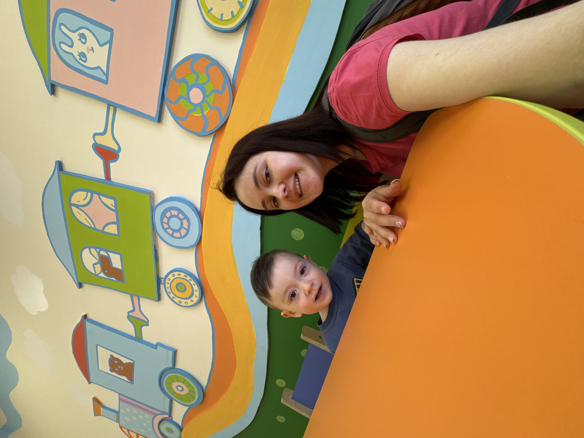 An adult woman and a young child are captured in a bright, cheerful indoor setting, likely in Izhevsk, Russia. The image shows both subjects smiling warmly and looking towards the camera.
The woman, positioned on the right side of the frame, has dark hair and wears a pink t-shirt and a dark backpack. Her left arm, featuring manicured nails, rests on a prominent, bright orange surface, which appears to be a table or part of a play structure.
Beside her, a young boy, likely a toddler, peeks over the orange surface. He has short dark hair and wears a dark blue long-sleeved shirt. He is also smiling at the camera, with his small hands gripping the edge of the orange surface.
The background features a colorful wall decoration, possibly a mural or 3D cutouts, depicting a stylized train. The train comprises multiple cars in pastel shades of pink, green, and light blue. Each train car has windows revealing cartoon animals, including a bunny, a bear, and another animal figure. The train's wheels are circular cutouts in various bright colors like blue, orange, yellow, and green. Wavy horizontal stripes in green, blue, orange, and yellow add to the playful scene, suggesting a landscape or a vibrant track. Some white cloud shapes are also visible on the wall.
The vibrant colors and child-centric decor suggest the setting is an indoor play area, daycare, or a similar child-friendly establishment. Both individuals display happy emotions, indicating a pleasant interaction or moment. There is no visible text in the image.