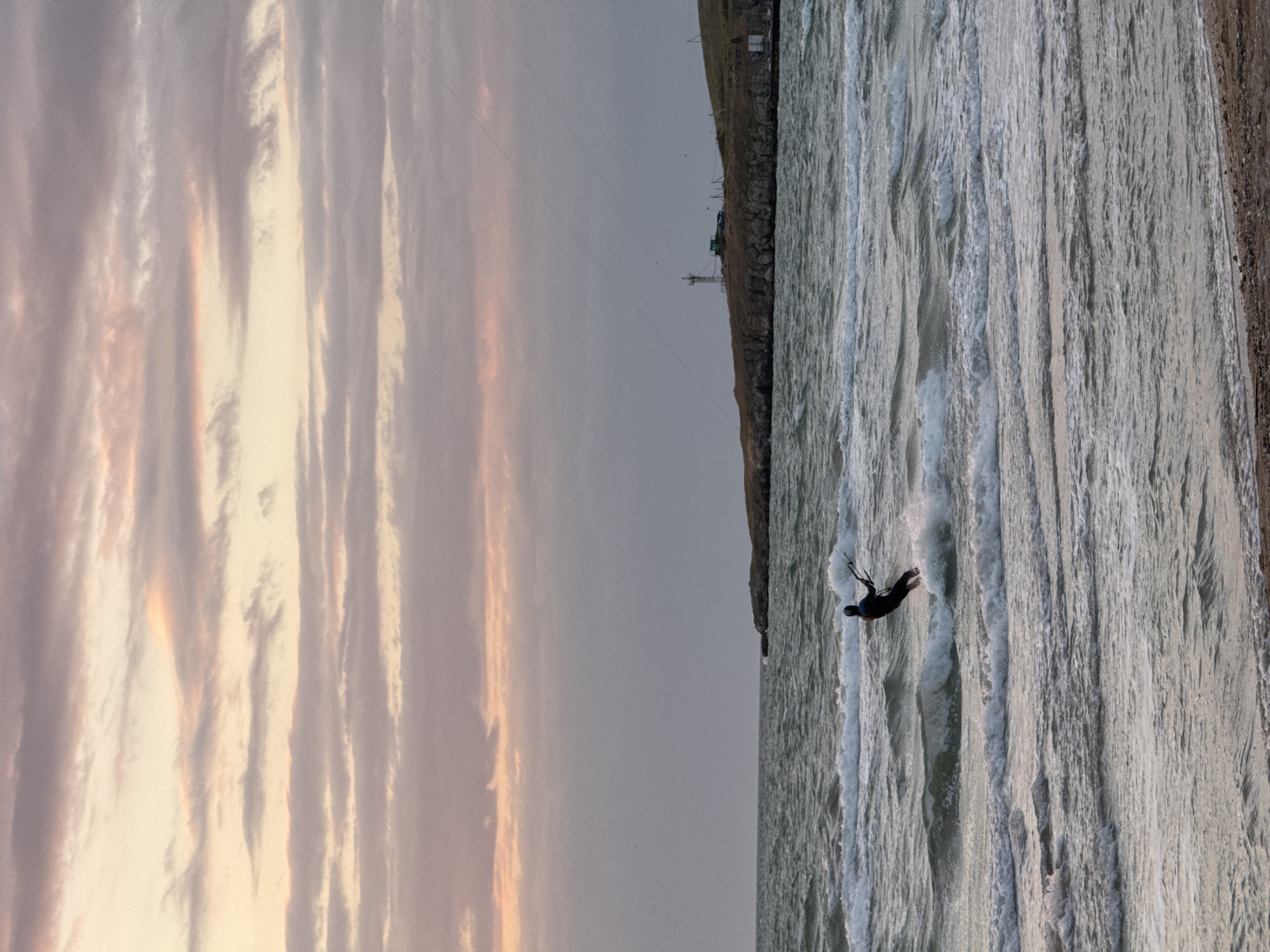 The image captures a kitesurfer in action off the coast of городское поселение Щелкино, Ukraine. The surfer, clad in a dark wetsuit, is in mid-maneuver on choppy water, with a kite line stretching diagonally upwards. The shoreline is visible to the right, transitioning from a rocky cliff face to a sandy beach. The sky dominates the left of the frame, featuring striated clouds tinged with hues of pink and orange from the setting sun. In the background, there are indistinct structures and possibly utility poles along the cliff top.