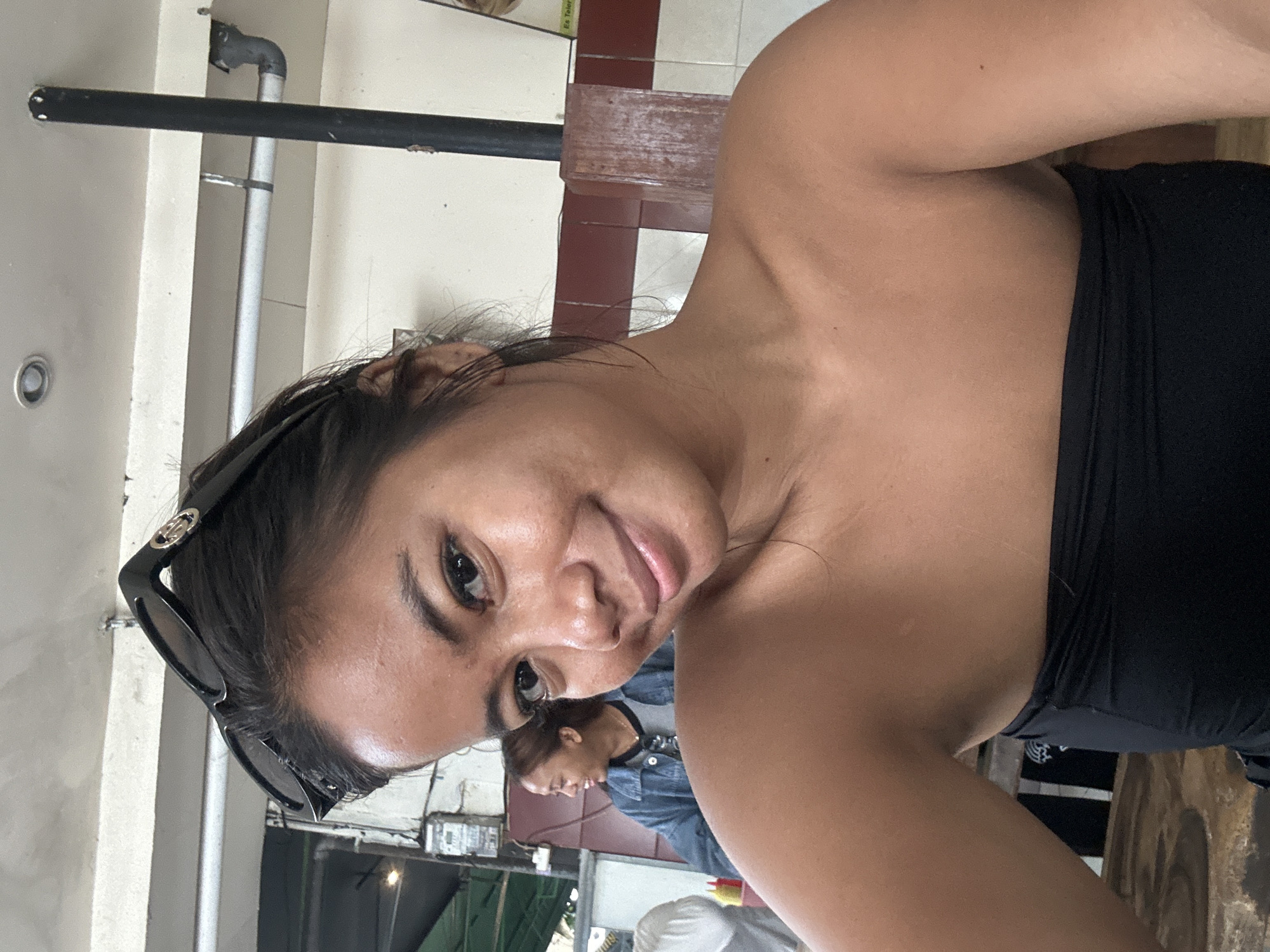 A young woman with sunglasses perched on her head takes a selfie in an outdoor seating area, possibly a restaurant, in Kuta, Indonesia. She is wearing a black strapless top. Behind her, another woman wearing a denim jacket is visible, smiling and looking to the left. The background includes a white wall with maroon trim, a wooden pillar, and some plumbing. The overall setting appears casual and public.