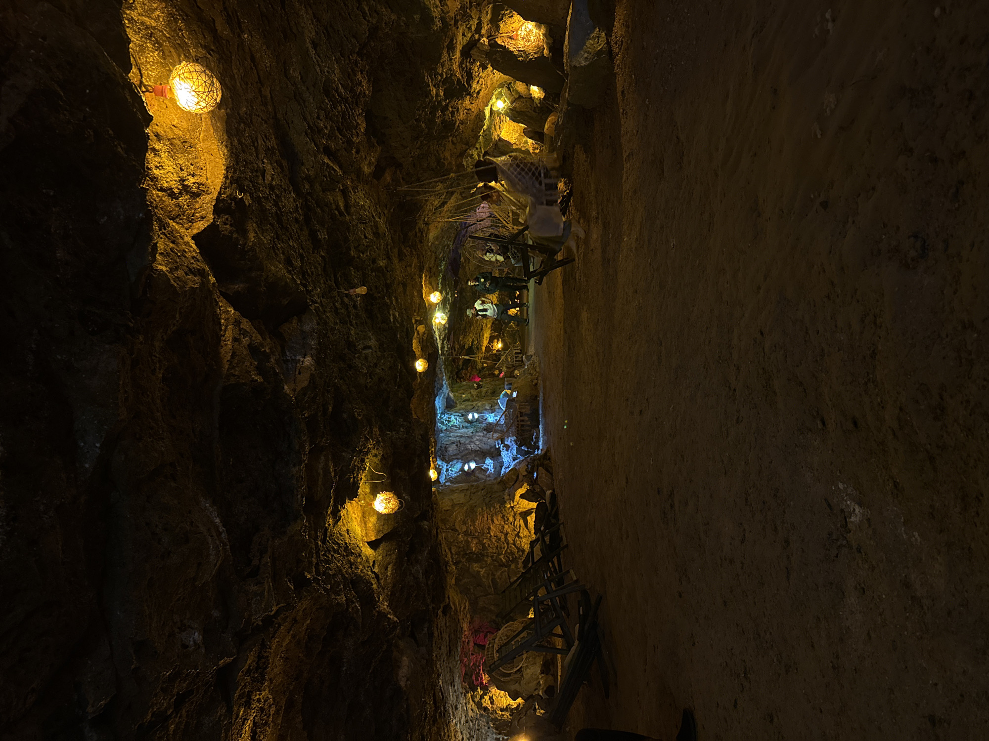 The image depicts the interior of a cave in Alaverdi, Armenia, illuminated by warm, decorative lighting. The cave's rough, natural rock ceiling and walls are visible, with several spherical, woven light fixtures hanging from above, casting a golden glow. In the foreground, the cave floor is covered in sand and dirt, with several empty, wooden lounge chairs arranged. Further into the cave, there are areas with more colorful lighting, including pink and blue hues, and some decorative elements that appear to be rock formations or water features. In the middle ground, several figures are present. To the left, three people are standing near a display, two of whom appear to be employees or guides in dark uniforms. To the right, a group of people are seated on a swing-like structure hanging from the cave ceiling, with another person standing nearby. The overall scene suggests an attraction or recreational space within the cave, designed for visitors to experience the unique environment.