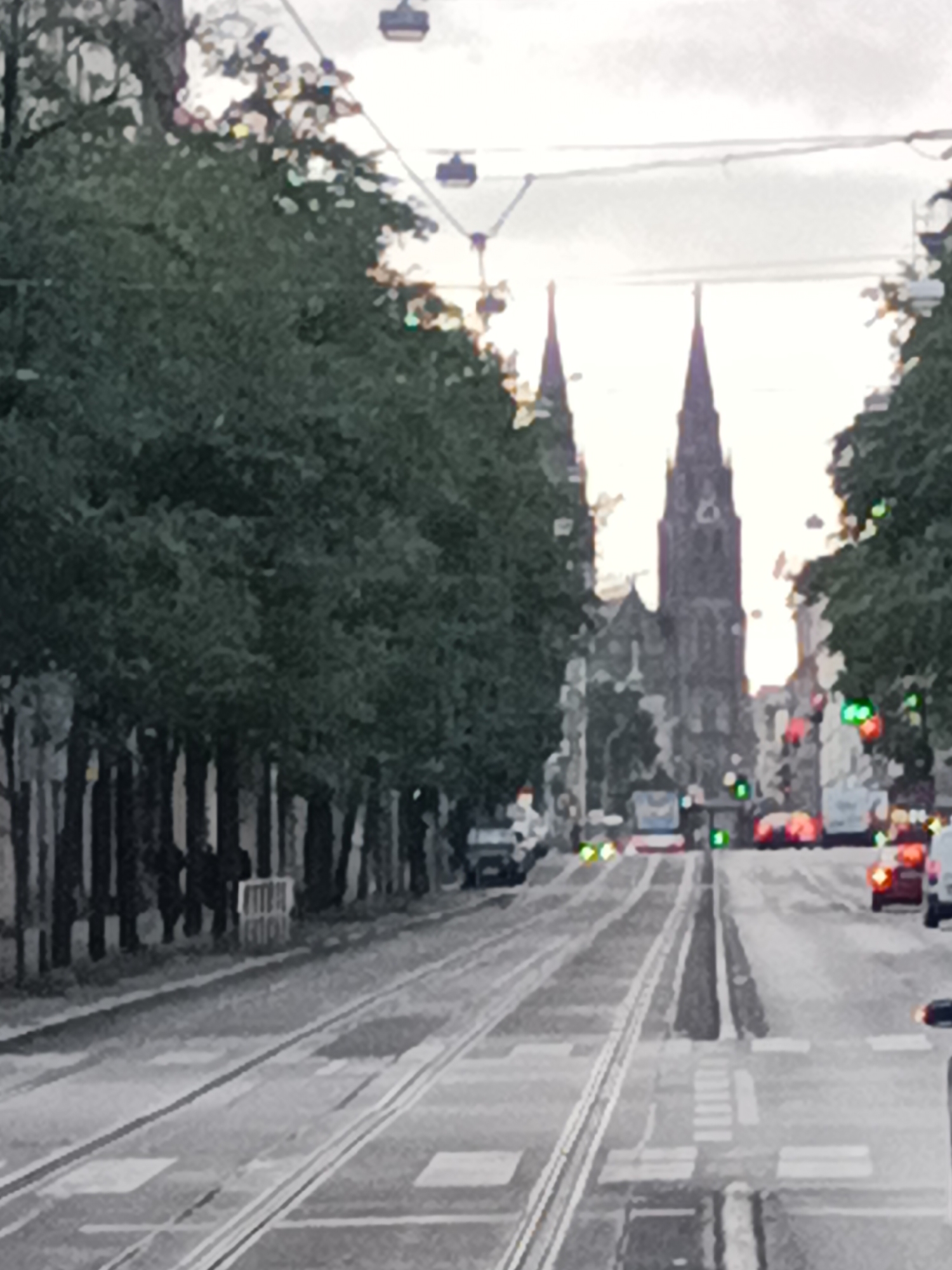 The image depicts a street scene in Prague, Czechia, with tram tracks running down the center of the road. On the left side of the street, a line of trees with dense foliage creates a canopy over the sidewalk. In the distance, two prominent spires of a Gothic-style church rise above the buildings and trees. The sky is overcast. Several vehicles, including cars and what appears to be a tram or bus, are visible on the road, along with traffic lights showing green. The overall impression is of a city street with significant architectural landmarks and active transportation.