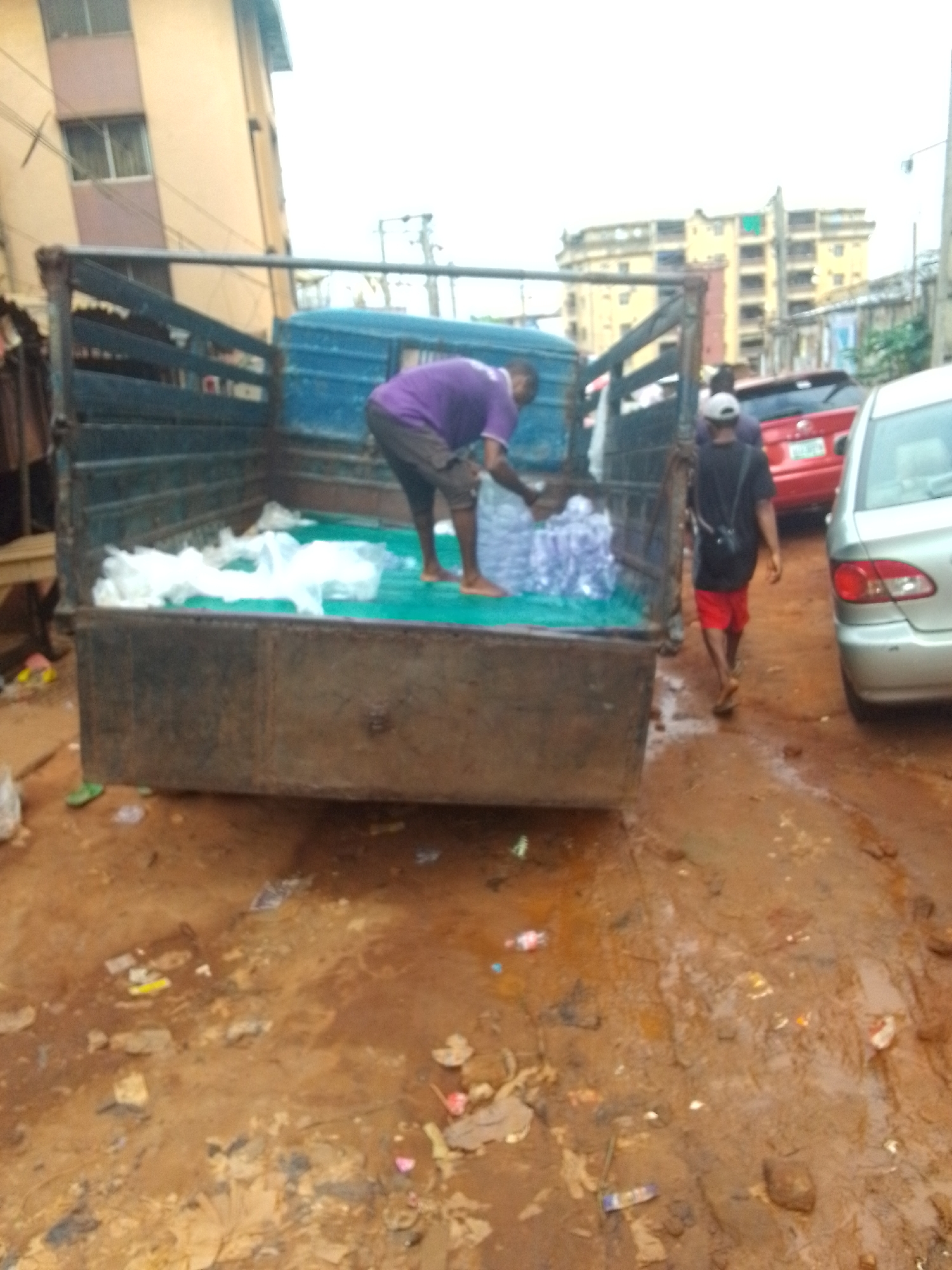 The image captures a scene in Onitsha, Nigeria, likely an urban or peri-urban area. A dark-colored truck is prominently featured, its bed filled with what appears to be bottled water. A man wearing a purple shirt and dark shorts stands in the truck, handling the water bottles. Another man, wearing a black shirt and red shorts, walks beside the truck.
Several buildings can be seen in the background, with one appearing to be multi-story. There are also cars nearby, including a red one and a silver one. The ground is muddy and littered with trash, suggesting a lack of sanitation. The image presents a scene of everyday life involving the transportation and distribution of water in a developing city.