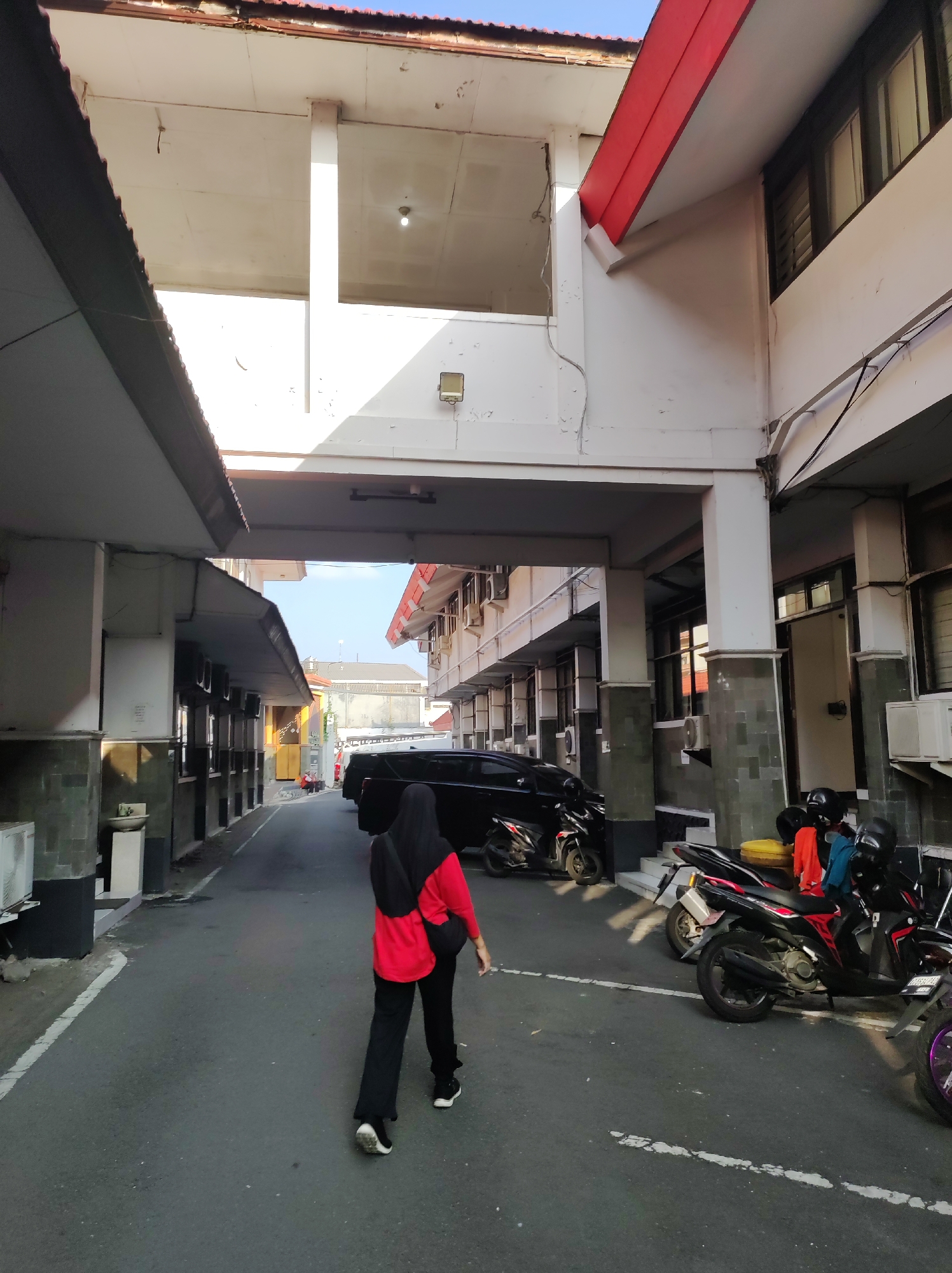 This is an outdoor, eye-level, full shot of a woman walking away from the camera down a narrow asphalt alleyway between two buildings. The woman is wearing a black hijab, a red long-sleeved shirt, and black pants. She is walking towards a black SUV parked in the middle of the alley. There are several motorcycles parked on the right side of the alley. The buildings on either side of the alley are white with dark trim and feature multiple windows. The buildings have overhead walkways supported by pillars. The sky is visible above the buildings and appears clear and blue. The scene is likely a commercial or institutional area, possibly within the city of Blitar, Indonesia, given its architectural style and the presence of motorcycles. The lighting suggests it is daytime, with sunlight casting shadows on the ground and buildings.