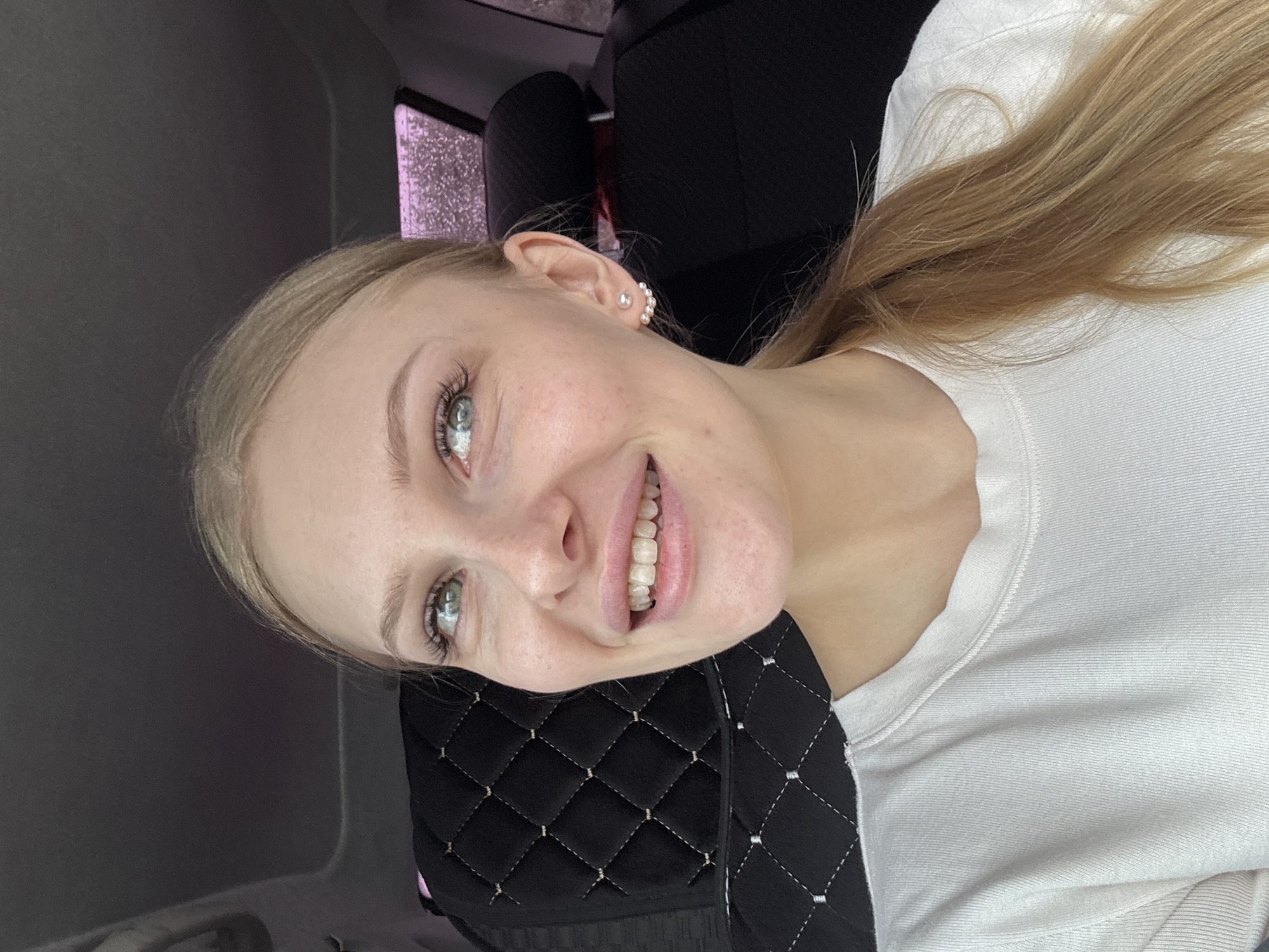 A young woman with light blonde hair pulled back in a ponytail is smiling and looking at the camera. She has fair skin and light blue eyes. She is wearing a white long-sleeved shirt and pearl earrings. She is seated in what appears to be a car, with a black quilted headrest behind her. The car interior is gray. Through the window, there's a blurred view that could be associated with the city of Perm, Russia, given the query context. The overall image has an informal, selfie-like quality.
