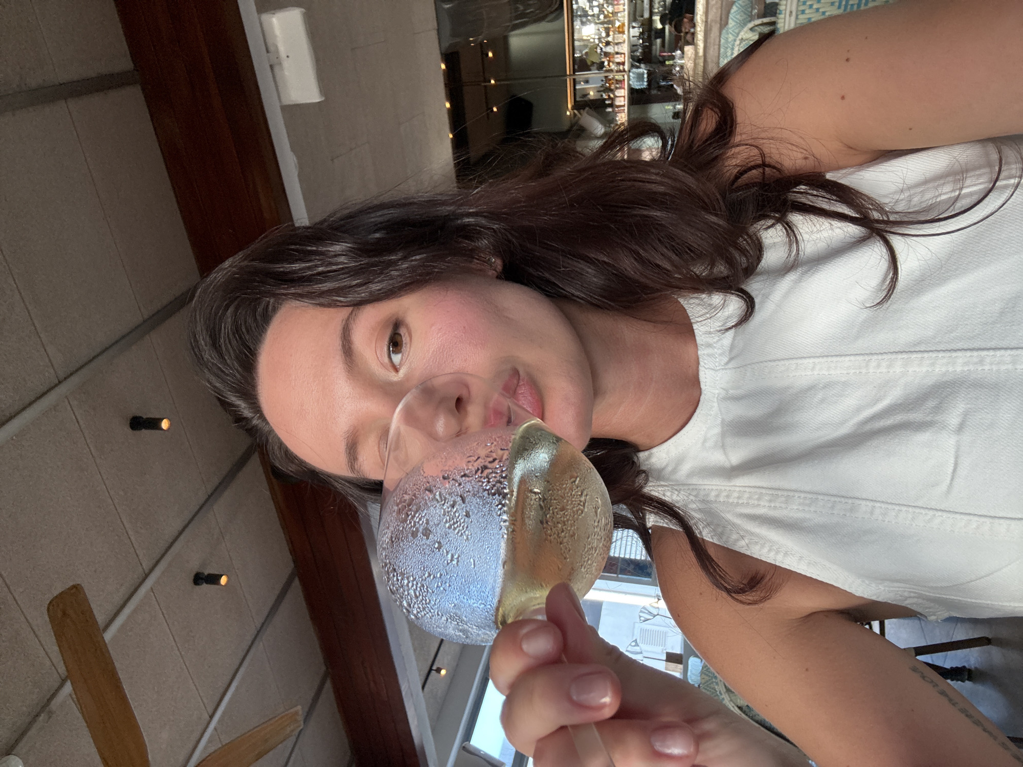 A woman with dark hair is taking a selfie indoors. She is holding a glass of white wine to her lips, her mouth puckered. She is wearing a white top. In the background, a bar with bottles on shelves is visible. The environment appears to be a restaurant or bar, possibly in Tel-Aviv, Israel, given the style of the ceiling fan and lighting fixtures, which are common in establishments there.