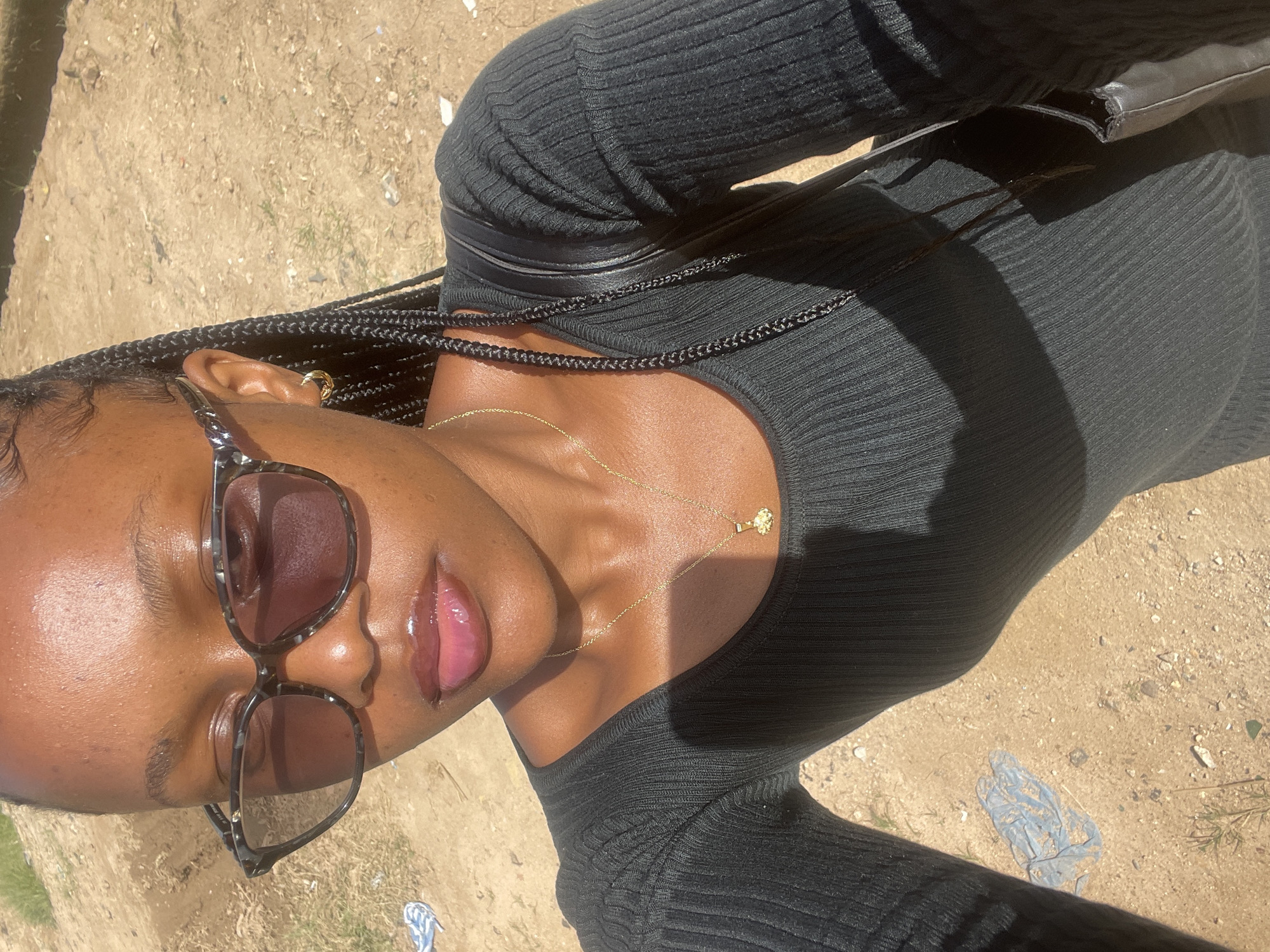 A young Black woman with dark braided hair and gold hoop earrings poses for a selfie outdoors. She is wearing dark sunglasses, a black long-sleeved ribbed dress, and a gold pendant necklace. A black purse is slung over her shoulder. The background appears to be a dusty, uneven ground with some sparse dry vegetation. The lighting suggests it is daytime. The location is identified as Jos, Nigeria.