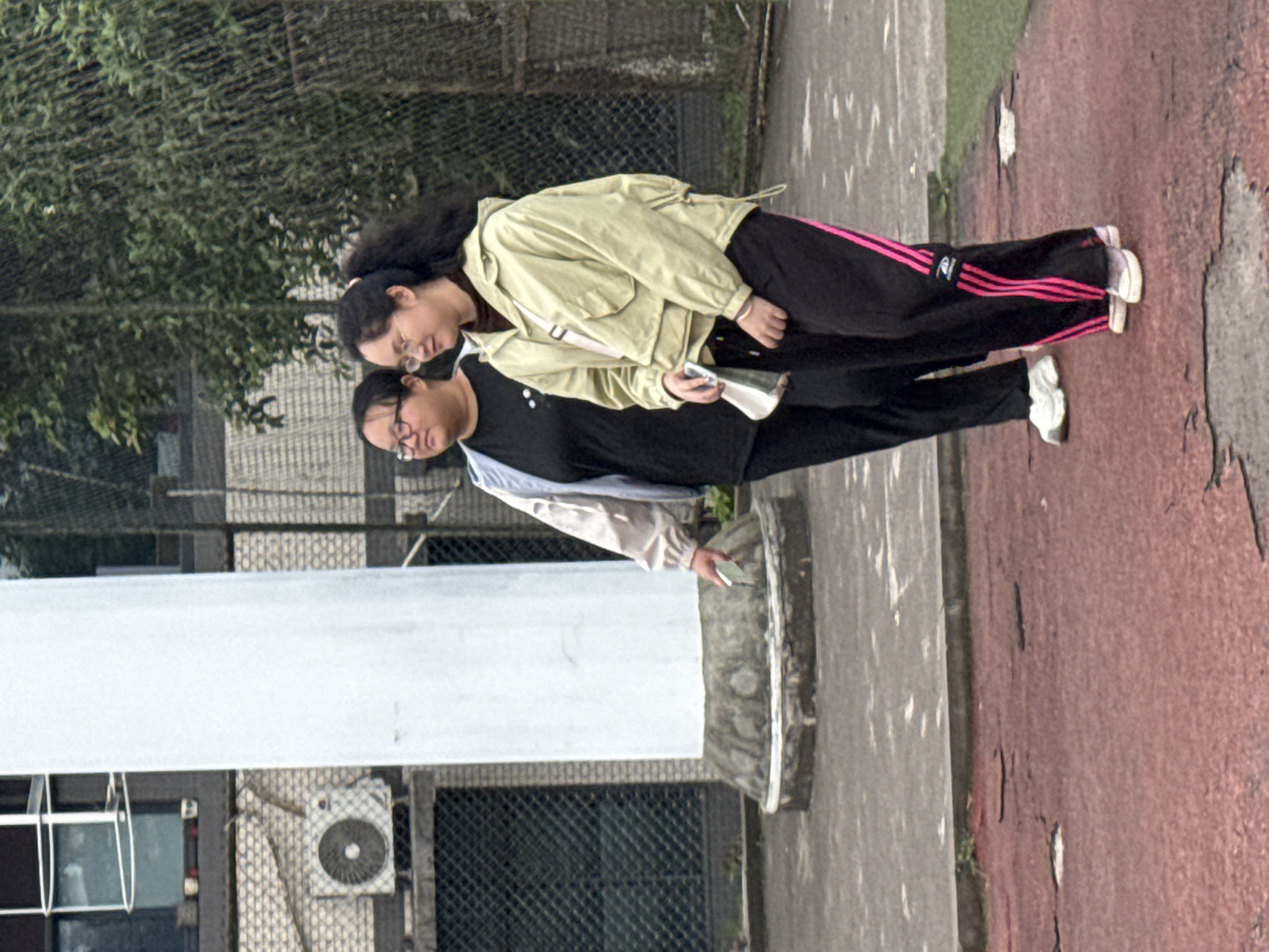 Two women are standing outdoors. One is wearing a black shirt, a light blue jacket, and glasses. The other is wearing a light yellow jacket and glasses, and black pants with pink stripes. They are holding phones and standing on pavement. In the background is a building, a fence, trees, and a basketball court. This image was likely taken in Jiangjin, China.