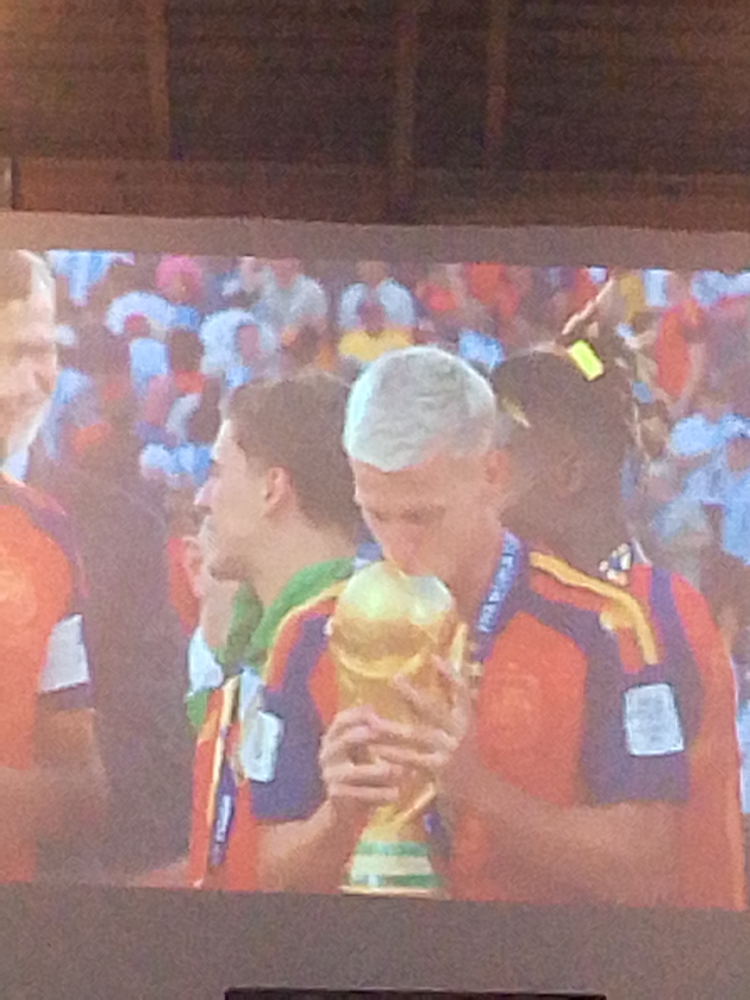 The image shows a projection or screen displaying a scene of people in what appears to be a celebration, likely a sporting event given the presence of a golden trophy. In the foreground, a man with short, light-colored hair is holding and kissing a large golden trophy. He is wearing a brightly colored jersey with horizontal stripes of purple, orange, and yellow accents. To his right, a person with dark skin and dark hair is partially visible, also wearing a similar jersey. To the left of the man holding the trophy, another man with dark hair is visible in profile, wearing a green and red striped garment. The background shows a crowd of people, with blurry figures and brightly colored clothing, suggesting a large gathering. The overall impression is of a moment of triumph and celebration. The image itself is blurry and pixelated, indicating it was likely taken from a distance or is a low-resolution digital capture. No specific details about Meunasahmee, Indonesia, are discernible from the image.
