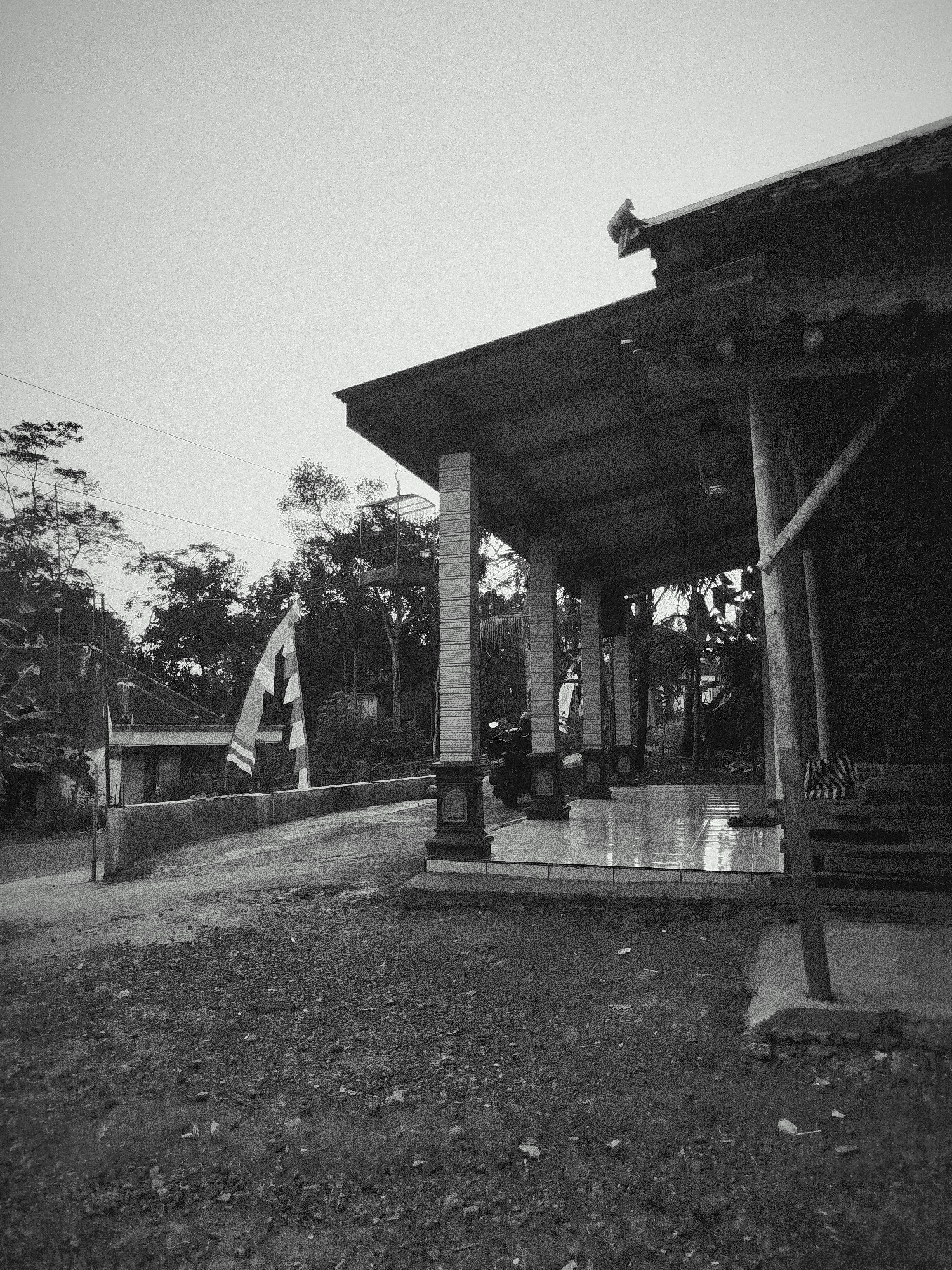 This black and white, grainy, wide-angle shot depicts an outdoor scene in Sewurejo, Indonesia, under an overcast sky. In the foreground, a rough, gravelly area leads to a paved path and a grey wall. Beyond the wall, a building with a dark, angled roof supported by pillars is visible. Decorative elements are present on the roof's edge. A flag with white and dark stripes is positioned on a pole near the building. In the background, numerous trees form a dense foliage. The scene appears to be a public or communal space, possibly near a temple or community center, with a motorcycle parked under the covered area. The overall impression is one of a quiet, possibly rural, Indonesian setting.
