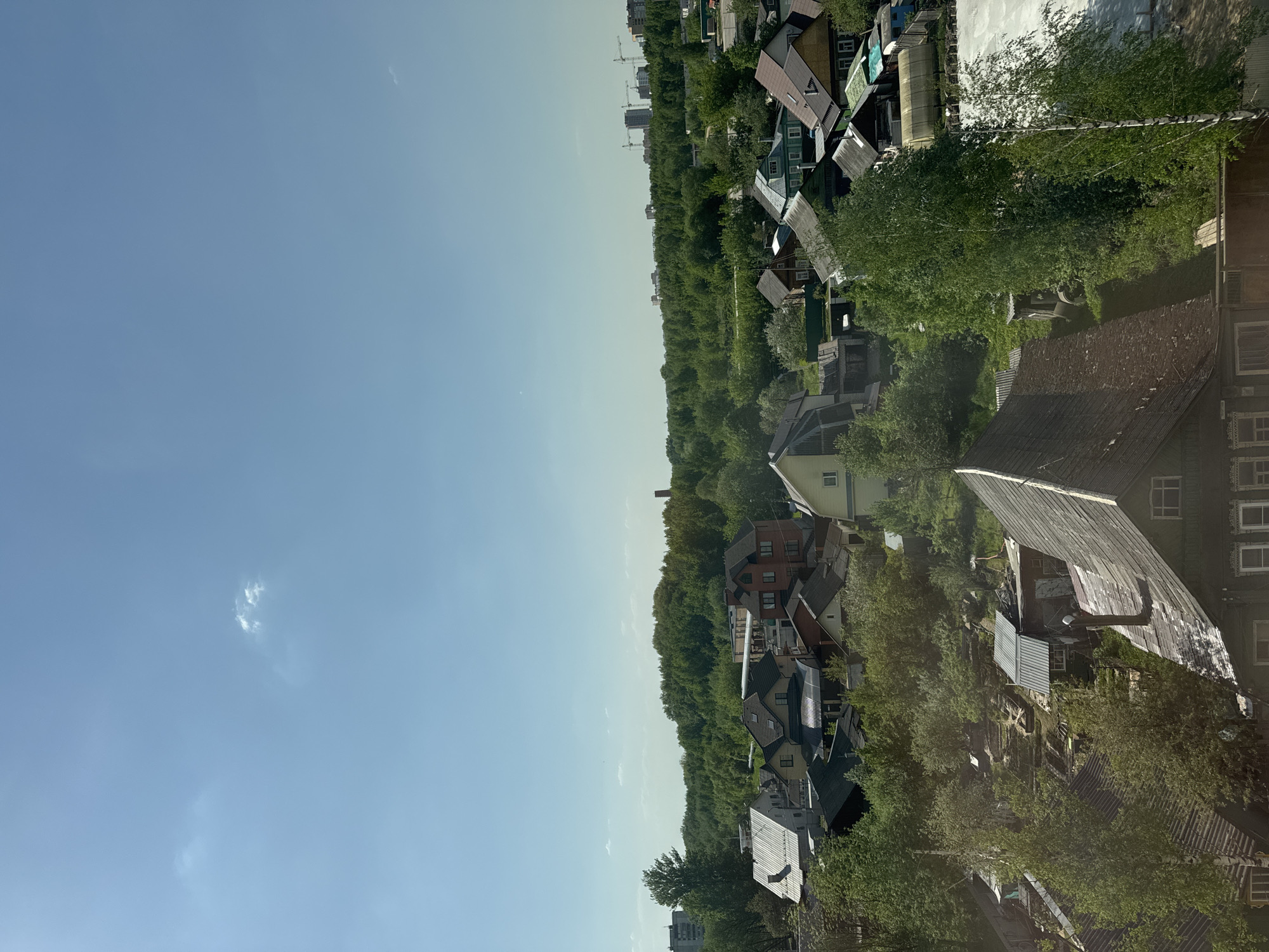 The image shows an aerial view of a neighborhood in Tver, Russia. The foreground is dominated by a blue sky with some small clouds. The right side of the image is filled with houses and trees. The houses are a mix of old wooden structures and more modern buildings. The trees are mostly green, suggesting that it is summertime. In the distance, there are a few taller buildings and construction cranes, indicating that the area is developing. The overall impression is of a peaceful, suburban neighborhood on a sunny day.