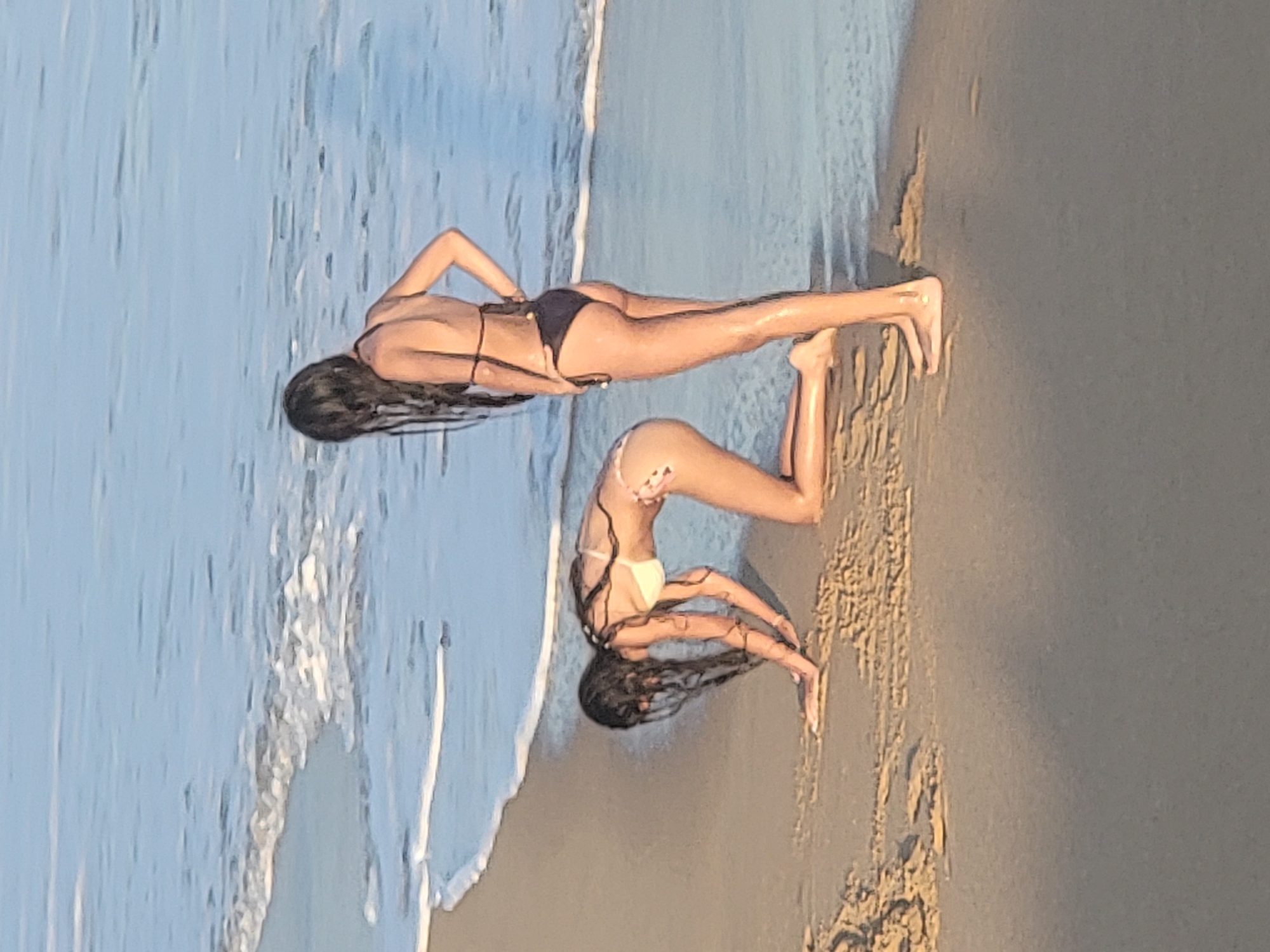 Two young women in bikinis are on a sandy beach near the water's edge in Quarteira, Portugal. One woman is kneeling on the sand with her hands outstretched. The other woman stands facing away from the camera, with one hand on her hip. The ocean is visible in the background, with small waves lapping at the shore. The lighting suggests it is either early morning or late afternoon.