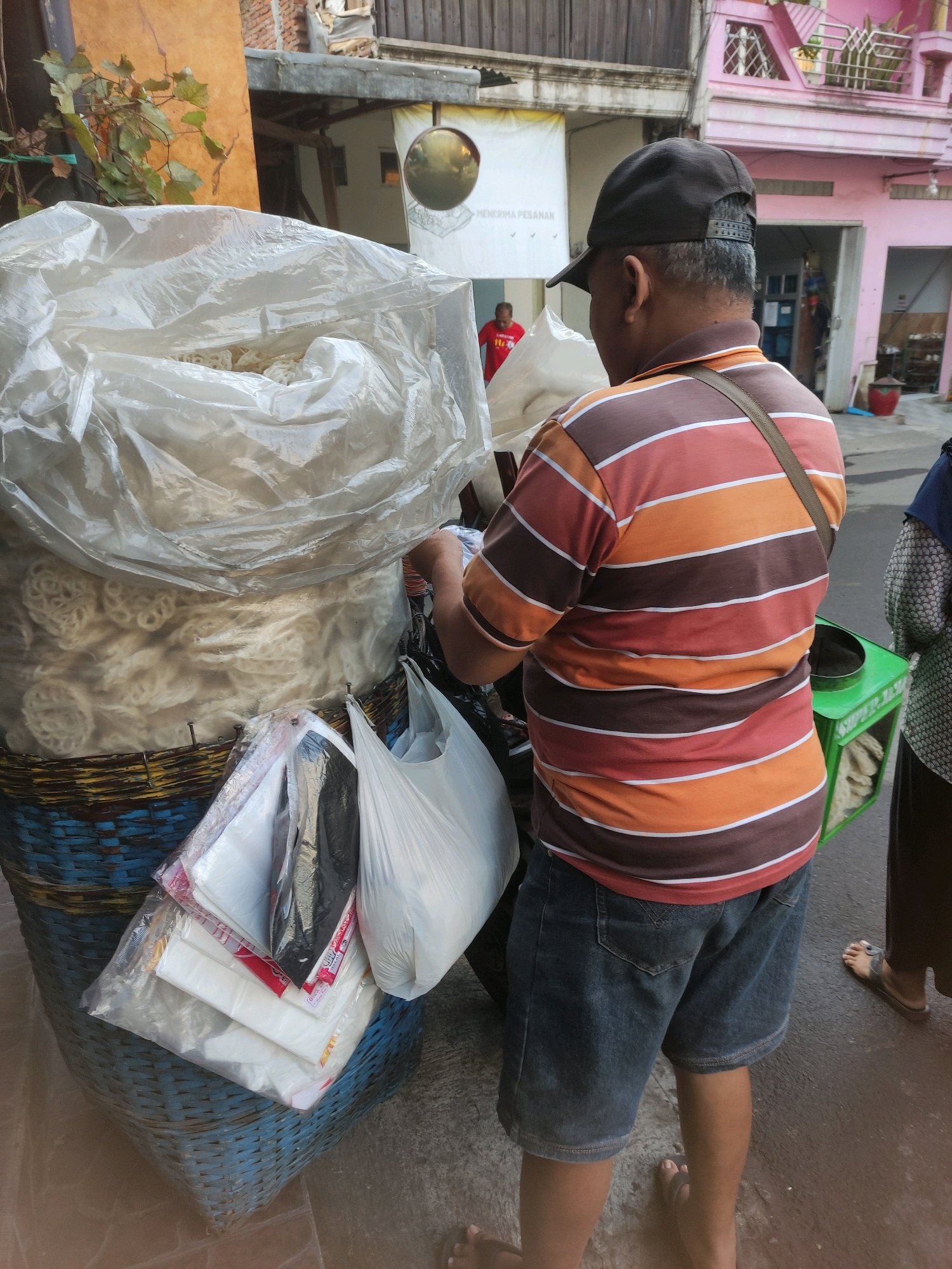 A man wearing a striped t-shirt and denim shorts stands on a street in Malang, Indonesia, next to a large basket filled with what appear to be crackers. The basket is covered with a plastic sheet, and several plastic bags are hanging from its side. In the background, a pink building with ornate windows is visible, along with other structures typical of a city environment. A woman in a headscarf is partially visible on the right side of the frame. The scene suggests a street vendor or market setting.