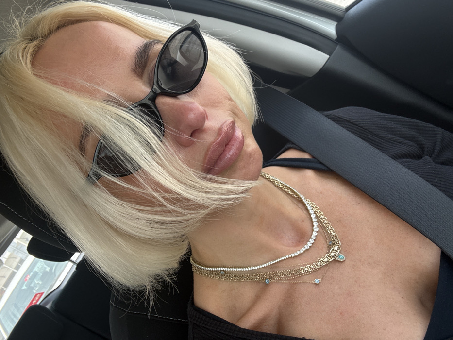 A close-up, high-angle selfie captures a person with short, platinum blonde hair, seated in a vehicle in Dubai, United Arab Emirates. The individual wears dark, cat-eye style sunglasses and a dark-colored top. A black seatbelt is fastened diagonally across the person's chest and right shoulder. The lips are slightly pursed and exhibit a glossy texture. Adorning the neck are multiple layered necklaces: a thick gold-toned chain, a choker-style necklace composed of numerous clear, faceted stones, and a delicate gold-toned chain featuring multiple small blue evil eye charms and a Hamsa hand pendant with a central blue stone. The vehicle's interior includes a dark headrest. Through a rear window, a portion of a white vehicle is partially visible in the background.