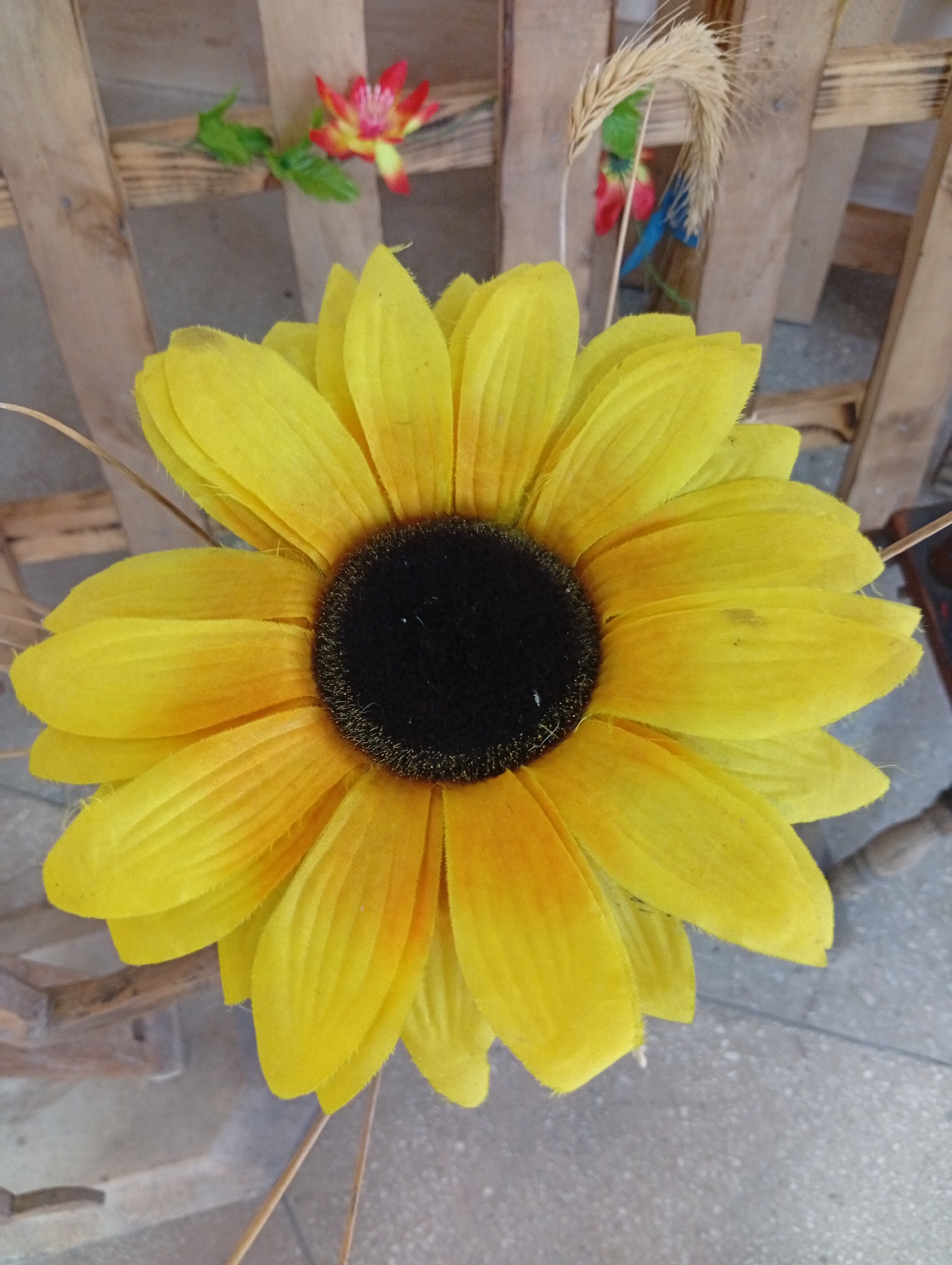 A bright yellow, artificial sunflower with a dark center is the main subject of the image, positioned centrally and filling most of the frame. Its petals are slightly uneven, with some showing slight discoloration or wear. In the background, a rustic wooden structure, possibly a fence or pallet, is visible. Behind the sunflower, to the left and right, are glimpses of small, artificial flowers in red and yellow, and a wispy, dried plant stalk. The ground beneath appears to be a gray, textured surface, possibly pavement or concrete. The image appears to be taken outdoors, with the lighting suggesting daylight. There are no people or animals visible in the scene. The location is specified as Kryŭlyanski Selski Savet, Belarus.