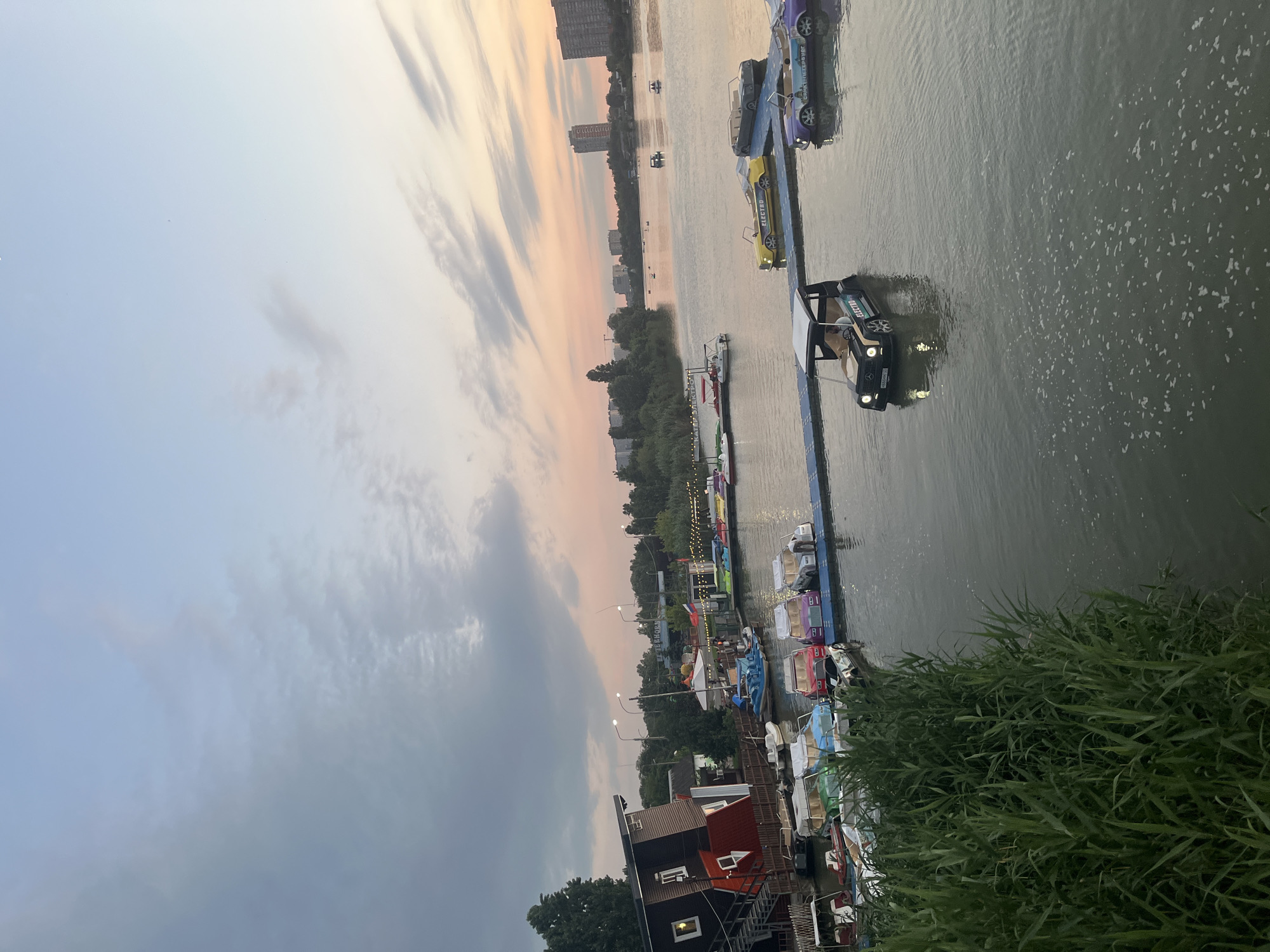 The image shows a waterfront scene at sunset in Krasnodar, Russia. The sky is a mix of pale blue and soft orange hues, with scattered clouds. In the foreground, tall green reeds line the edge of a body of water. Several boats are docked along a pier. These include pedal boats shaped like cars, with some yellow, purple, and white models visible. One black car-shaped pedal boat is actively in the water, with a person inside. Further back, more boats are moored, including a small blue motorboat and several white pedal boats. On the shore, there is a building that appears to be tilted or intentionally designed to look so, with a red roof and dark siding. Other structures, including a tent and more buildings, are visible along the waterfront. In the distance, trees and apartment buildings line the opposite bank of the water. A few other boats are scattered across the water, and some people can be seen in the distance. The overall atmosphere is calm and recreational.