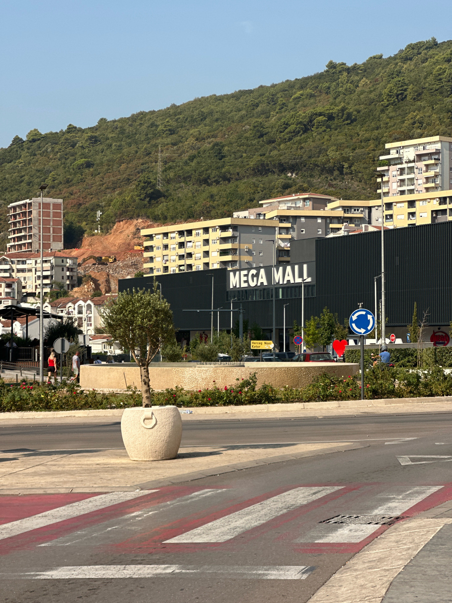 The image depicts a commercial area in Budva, Montenegro, with the "MEGA MALL" prominently displayed on a dark, modern building. In the foreground, a pedestrian crosswalk with red and white stripes leads across an asphalt road. A circular planter with a small tree and white decorative stones sits to the right of the crosswalk. Behind the planter, a low wall adorned with textured patterns and flowering bushes separates the road from the mall's entrance area.
The mall's facade is dark and features large, white lettering spelling out "MEGA MALL." Several streetlights and signage are visible in front of the building. To the left of the mall, other buildings with varying architectural styles, including apartment complexes and unfinished construction, are visible.
In the background, a steep, tree-covered hill rises behind the buildings. Construction activity, including heavy machinery like an excavator, is evident on a section of the hillside. The sky is clear and blue, indicating a sunny day. A few people are seen walking on a sidewalk near the mall, and a person on a scooter is visible in the distance. Road signs, including a blue circular sign with white arrows indicating a roundabout and a red circular "stop" sign, are present. The overall scene suggests a developing urban environment where commercial and residential development meets the natural landscape.