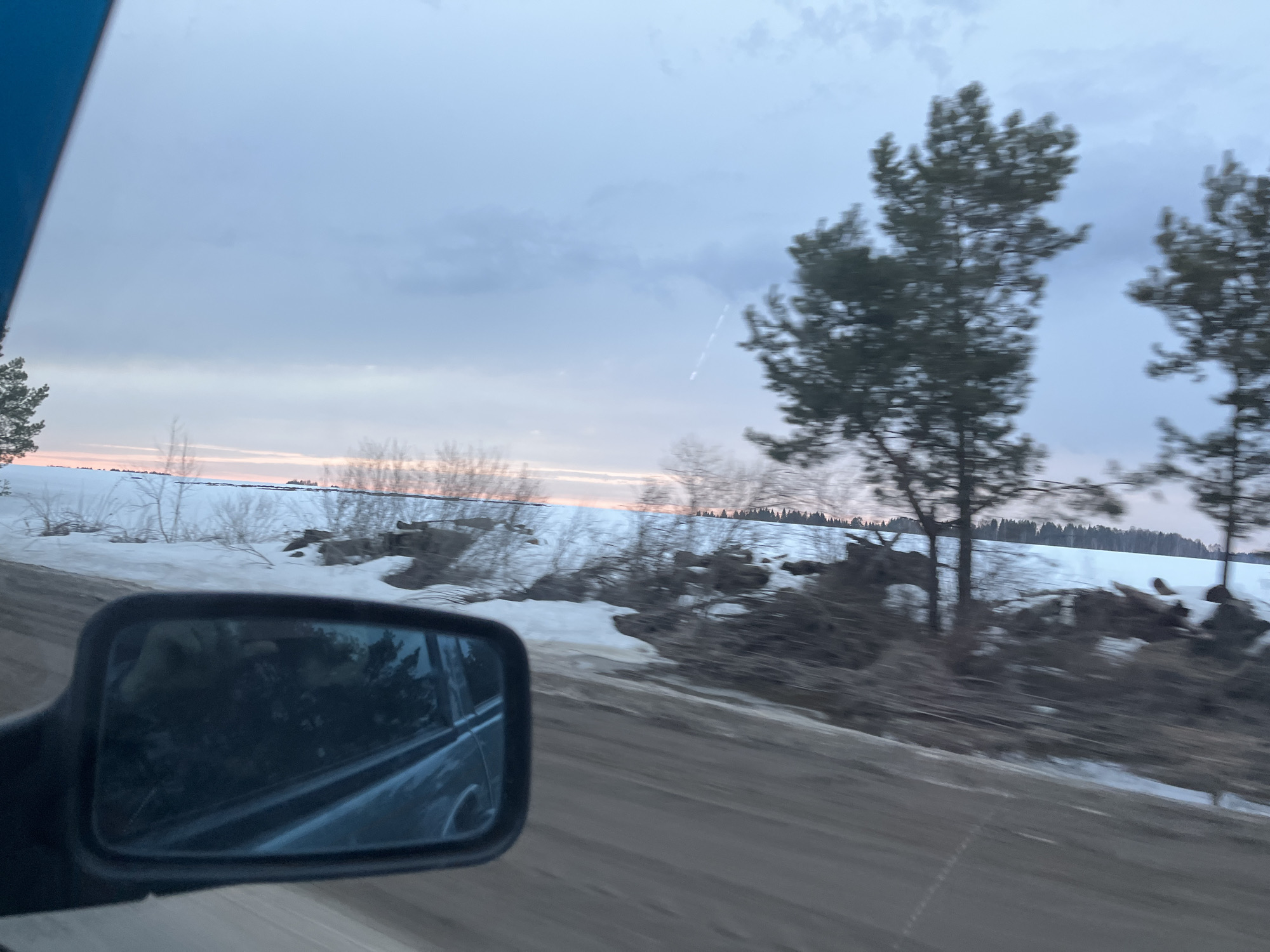 The photo depicts a blurred roadside landscape in Chumna, Russia, taken from a moving vehicle. Patches of snow cover the ground, and bare trees and shrubs populate the foreground and middle ground. A frozen lake or field stretches into the distance, with a faint orange glow visible on the horizon under a cloudy sky. In the lower left corner, a side mirror reflects an interior view of the vehicle. The road surface is asphalt.