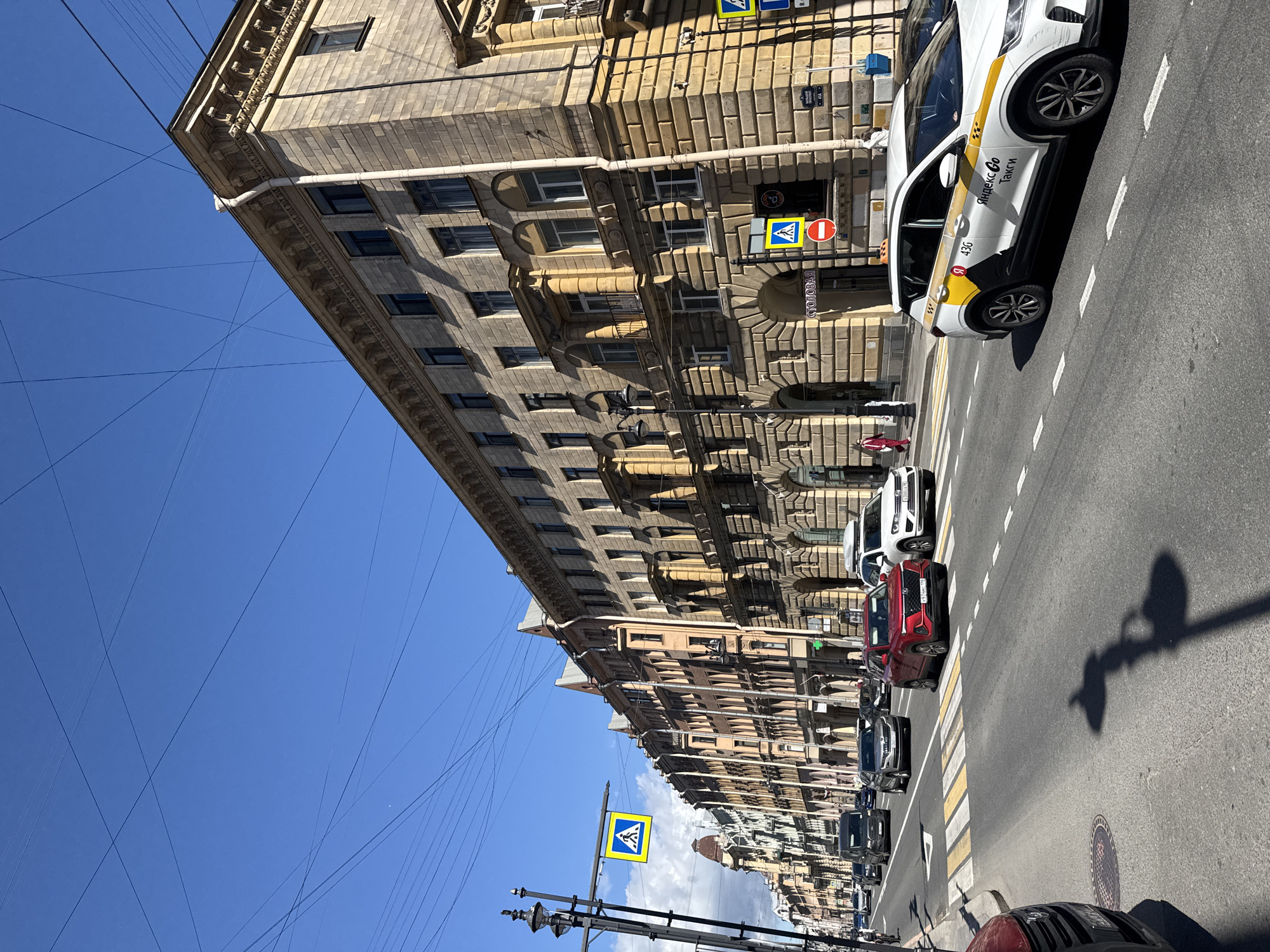 In this sun-drenched urban scene, a wide street in St. Petersburg, Russia, is lined with ornate, multi-story buildings. The architecture features classical elements like columns and arched doorways, with facades constructed from light-colored stone. Overhead, a network of dark electrical wires crisscrosses the bright blue sky.
The street itself is bustling with traffic. Several cars, including a red SUV, a silver sedan, and a white taxi with "Яндекс Go Такси" branding, are captured in motion. A pedestrian crossing with yellow and white stripes is prominent, and a yellow pedestrian crossing sign with a blue pictogram is visible on the left. A red traffic signal is also present.
On the sidewalks, a few pedestrians can be seen, including a person in a bright red suit walking away from the camera. Shadows of lampposts stretch across the asphalt, indicating a clear, sunny day. The overall impression is a vibrant city street on a typical day.