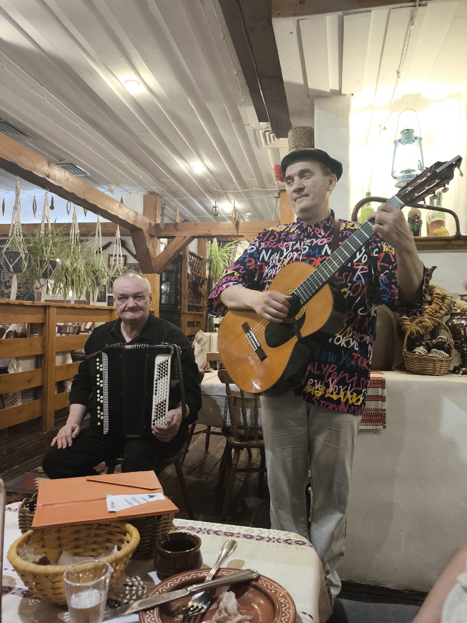 Inside a restaurant, two musicians are performing. One man is sitting and playing an accordion. He is wearing a black shirt and pants. The other man is standing and playing a guitar. He is wearing a hat, a brightly colored shirt with words on it, and light-colored pants. There is a table in front of them with food, drinks, and an orange folder. In the background, there are wooden beams, plants, and a decorative shelf with various items. The location is likely in Moscow, Russia based on the restaurant's architectural style and decorations that are consistent with Russian folk themes.