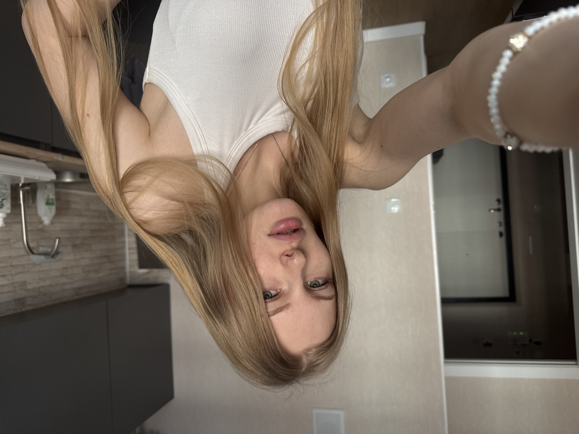 A young woman with long blonde hair is taking a selfie in what appears to be an apartment. She is wearing a white tank top and a beaded bracelet. The background shows a bathroom with a dark gray vanity, tiled wall, sink, and a towel hook. The other side of the room features a light-colored wall and a partially visible doorway or window with a dark frame. The city of Perm, Russia, is not ascertainable from the content of the image.