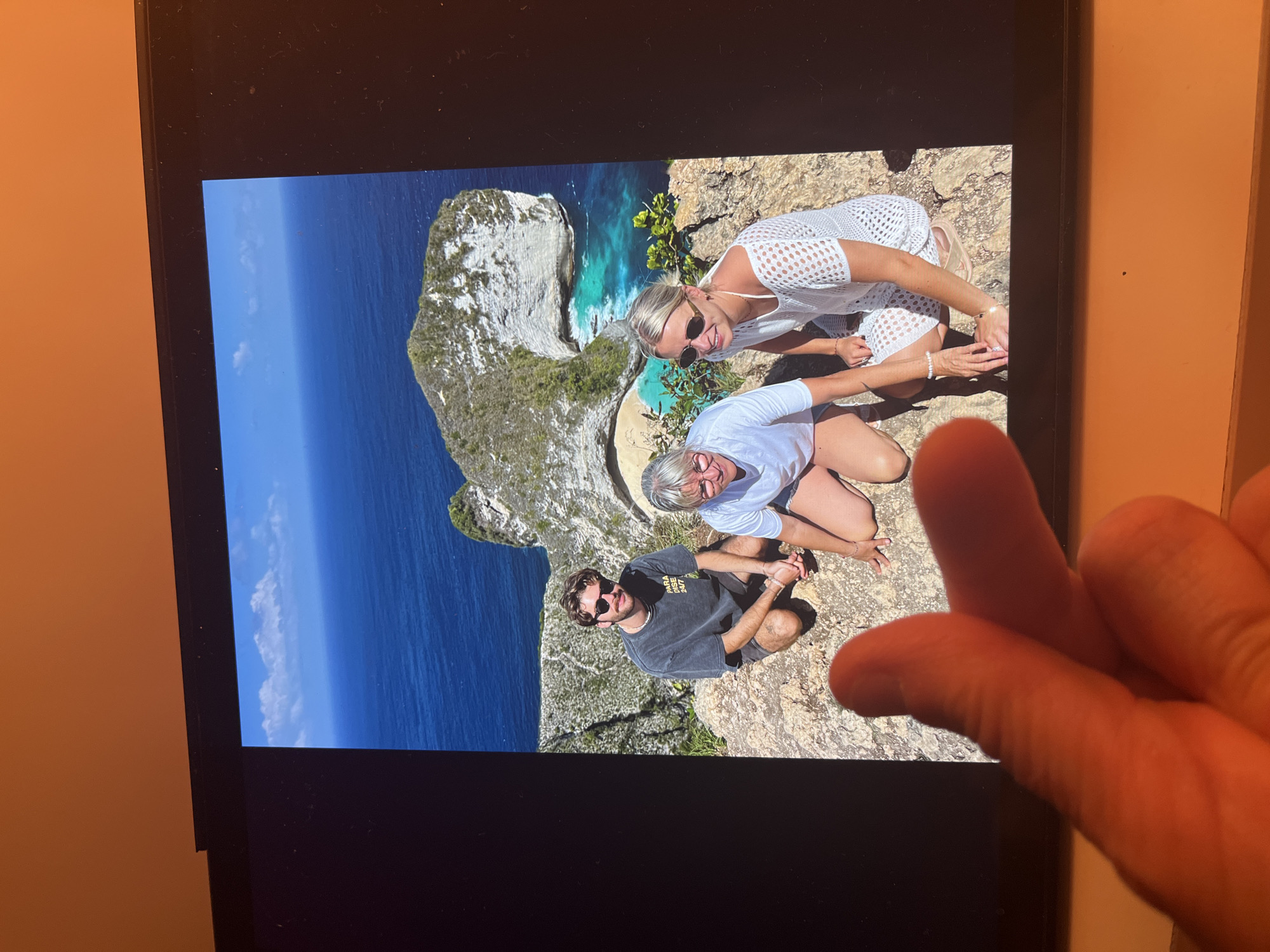 The image, captured in Chambœuf, France, displays a digital screen featuring a photograph of three individuals on a cliffside. The photograph shows a man and two women crouching on a rocky surface. The man, positioned on the left, wears a dark t-shirt, shorts, sunglasses, and a necklace. The central woman wears a white t-shirt, light shorts, and glasses. The woman on the right wears a white open-knit dress and sunglasses. All three are smiling and looking towards the viewer. The background of the displayed photograph presents a dramatic coastal scene with a distinctive white cliff formation jutting into the ocean, a small sandy beach with clear turquoise water, and deep blue sea extending to the horizon under a bright sky with scattered white clouds. The digital screen itself has a black bezel. In the immediate foreground of the overall image, a human hand with two fingers, illuminated by a warm, reddish light, partially obscures the lower-left portion of the screen. The surrounding environment, consistent with its location in Chambœuf, France, features warm orange-toned walls visible above and below the screen.
