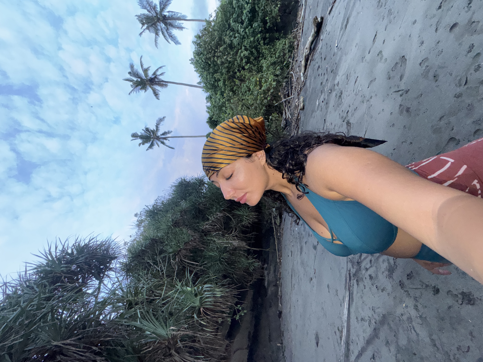 A young woman is the focal point of the image, set against a backdrop of a beach. She wears a blue swimsuit top and a tiger-striped bandana. Her head is tilted downwards.
The location is a beach in Selemadeg Timur, Indonesia. Tall palm trees are visible in the upper portion of the frame against a partly cloudy sky. Lush green vegetation edges the beach, meeting a dark sandy shore.