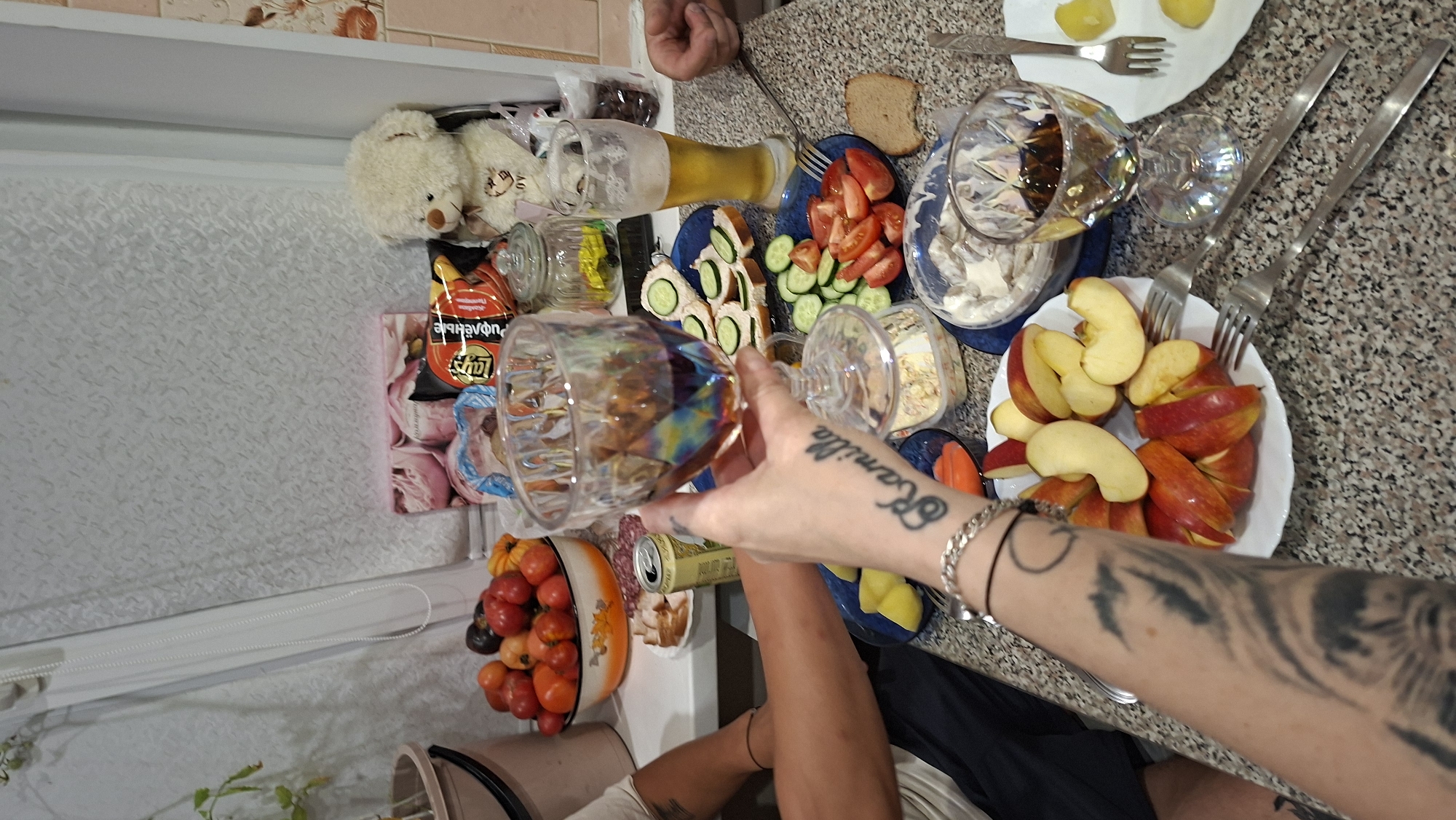 The image depicts a close-up view of a table laden with food and drinks, suggesting a meal is in progress. A person's arm, adorned with tattoos and a bracelet, is prominently featured holding a stemmed glass filled with a dark liquid, possibly wine. The arm extends from the left side of the frame, with the tattoos showing abstract designs and the name "Kamilla" in cursive script.
On the table, several plates contain various food items: sliced apples, a salad with cucumbers and tomatoes, what appears to be herring or a similar fish dish, and sandwiches with cucumber slices. A bowl of mixed fruits, including tomatoes and other small, round fruits, is visible in the background. A can, possibly of soda or beer, sits near a plate with slices of cured meat. A glass of beer, with a frothy head, is also present. In the far background, a white teddy bear is seated, partially obscured by other items. A clear glass jar filled with a yellow substance and a bag of chips are also visible.
The table surface has a speckled, granite-like pattern. The background suggests an indoor setting, possibly a kitchen or dining area, with a window covered by a white blind. The lighting appears to be artificial, casting a warm glow on the scene.
While the image is rich in detail about the meal and the objects present, it does not provide any direct information to place the scene specifically in Myadzyel, Belarus. The overall impression is one of a casual gathering or mealtime.
