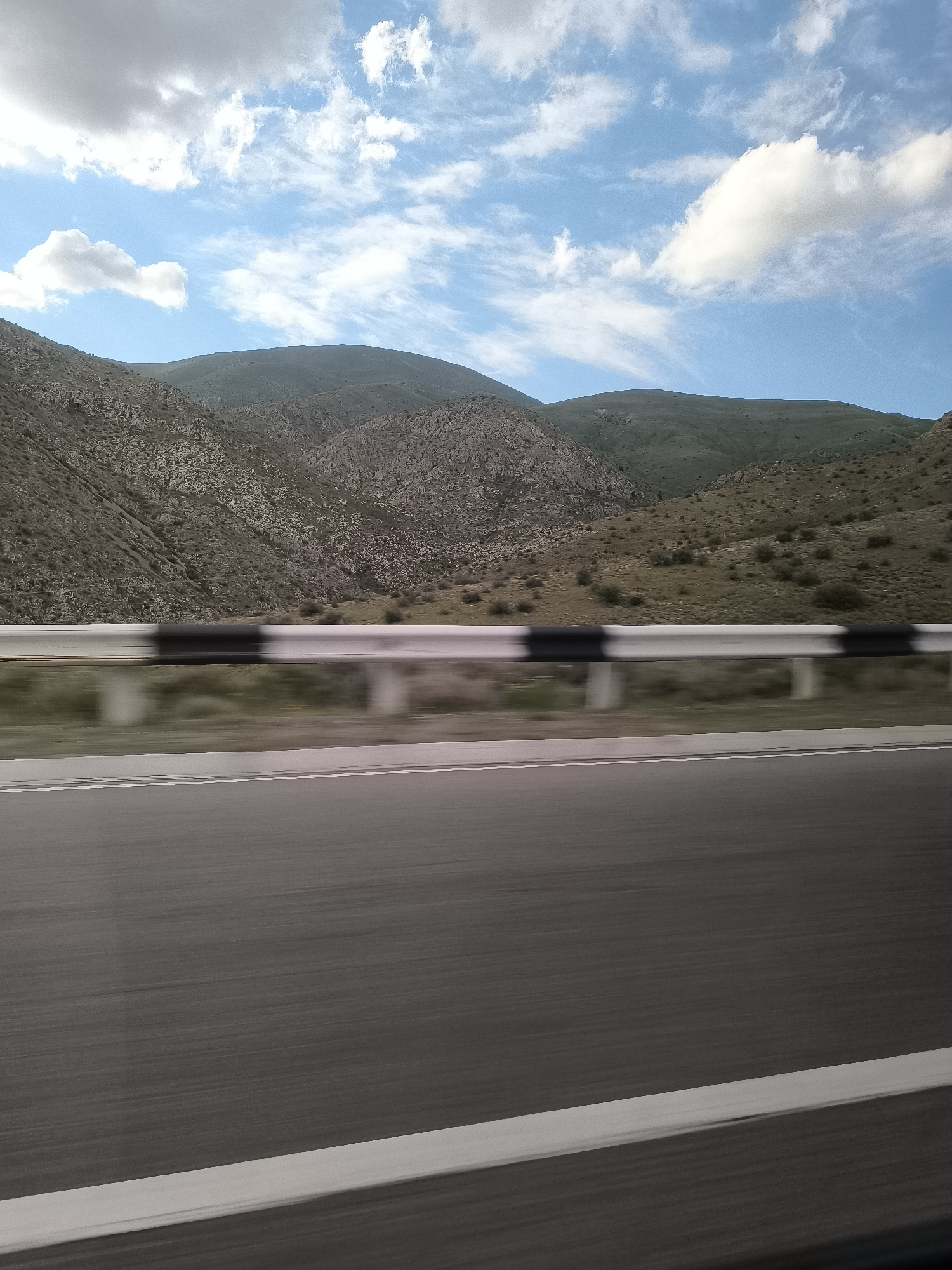 The image presents a dynamic view from a moving vehicle traversing a road through the mountainous landscape of Tigranashen, Armenia. In the foreground, a multi-lane asphalt road stretches ahead, with its surface and a prominent white solid line on the right shoulder appearing blurred due to motion.
A metal guardrail, painted with distinct black and white sections, runs parallel to the road, also exhibiting motion blur, emphasizing the speed of travel. Beyond the guardrail, the terrain rises into rugged mountains and hills, characterized by rocky outcrops and sparse, dry vegetation, predominantly in shades of brown and muted green. The contours of the mountains suggest a varied and textured topography.
The upper portion of the frame is filled with a bright blue sky, adorned with scattered white clouds of varying shapes and sizes, from fluffy cumulus to more spread-out, wispy formations. The clear sky and strong lighting indicate it is daytime, likely mid-morning or afternoon. No people or visible text are present in the image. The scene conveys a sense of journey and the vastness of the natural environment.