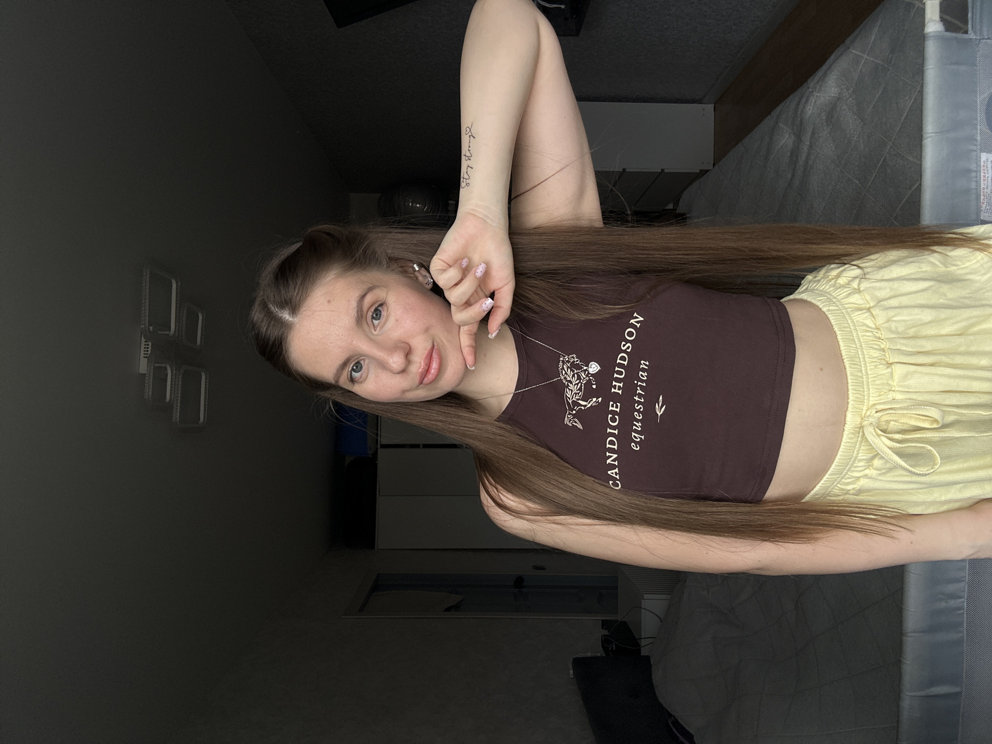 A young woman is posing for a selfie indoors. She has long, straight brown hair with a partial updo, a brown crop top with the text "CANDICE HUDSON equestrian," and light yellow drawstring pants. She is wearing a necklace and has a tattoo on her forearm that says "Stay strong." The background includes a dark wall, a wardrobe, a bed with a grey quilted cover, and what appears to be a baby cot. There is no indication in the image that the location is Klin, Russia.