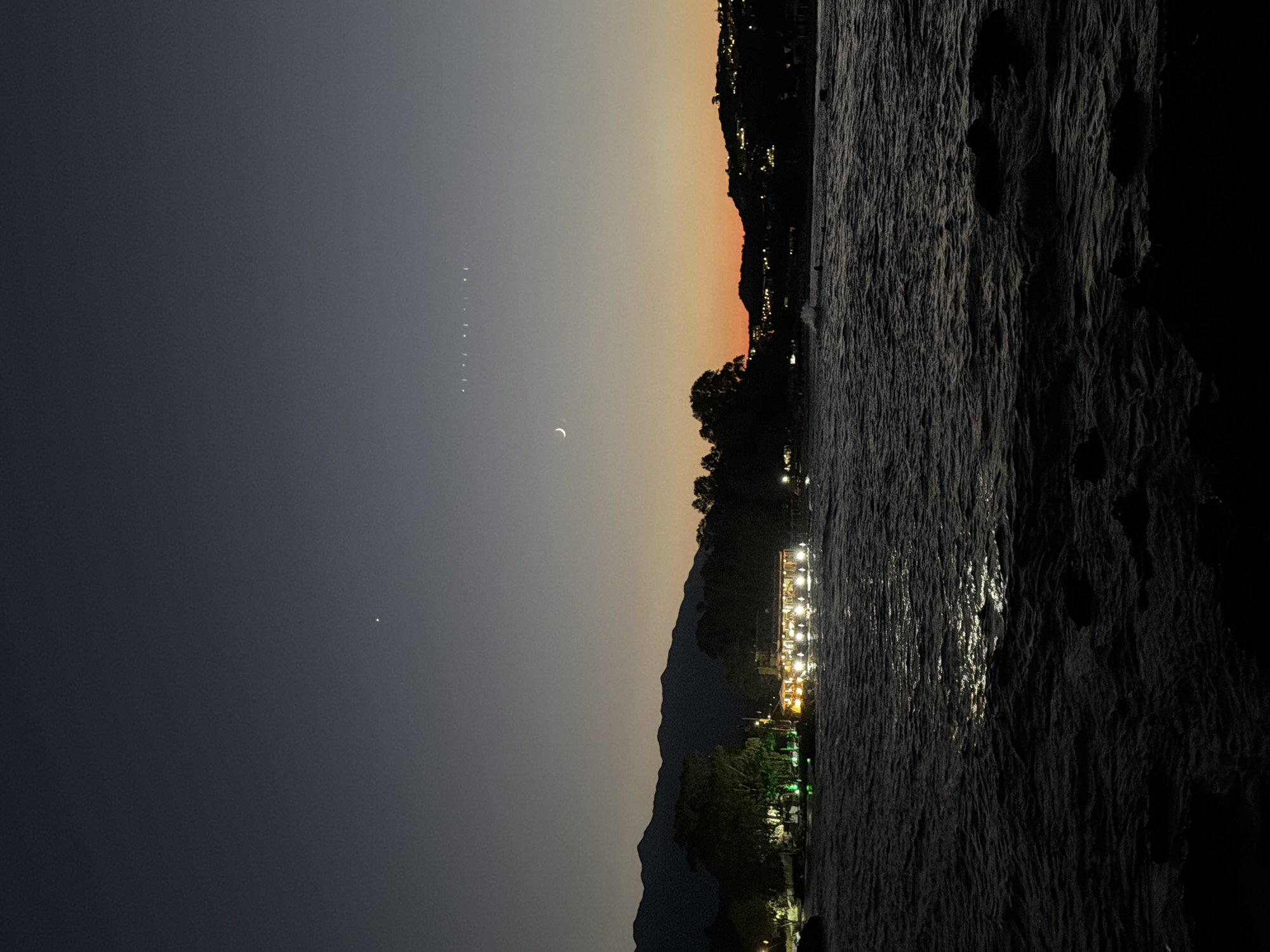 This vertical, medium-range shot captures a twilight scene over the Aegean Sea near Georgioupoli, Greece. The sky transitions from a deep, dusky blue at the top to a warm, orange glow near the horizon. A thin crescent moon is visible in the upper half of the frame. Below the sky, a dark silhouette of mountains stretches across the background. In the mid-ground, a line of trees and buildings are illuminated with warm lights, suggesting a waterfront establishment. The foreground is dominated by the dark, rippling surface of the sea, with rocks visible at the bottom edge of the frame. The overall mood is serene and tranquil, characteristic of a peaceful evening by the sea.