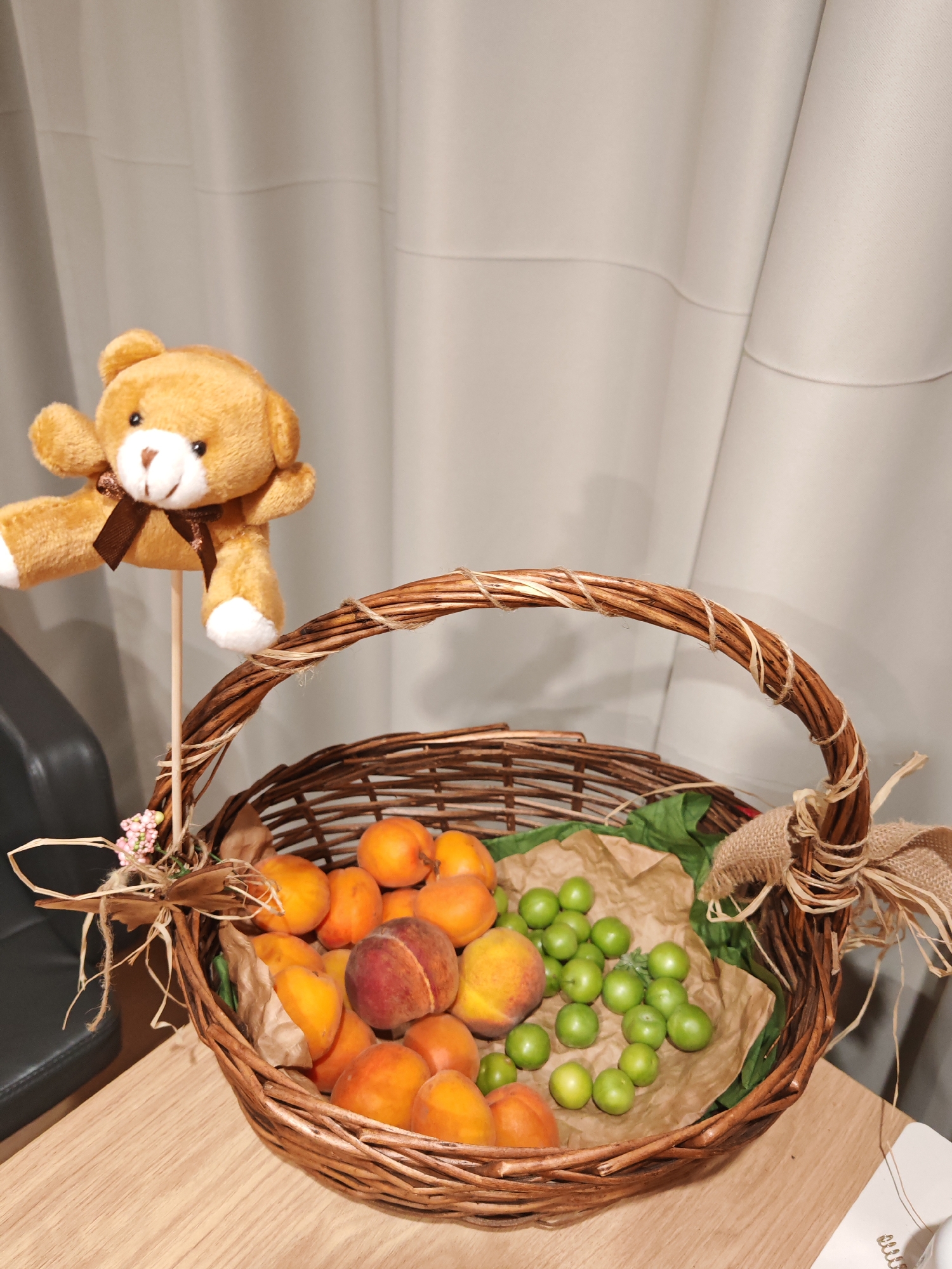 A teddy bear is attached to a stick and placed in a woven basket filled with fruits. The basket is made of brown twigs and has a handle wrapped with twine. Inside the basket are peaches, apricots, and green plums. The basket sits on a wooden table. In the background, a beige curtain is visible. A black chair is partially seen to the left of the basket. The scene is indoors. The image does not contain any specific elements that would allow for the localization of the city of Plovdiv, Bulgaria.