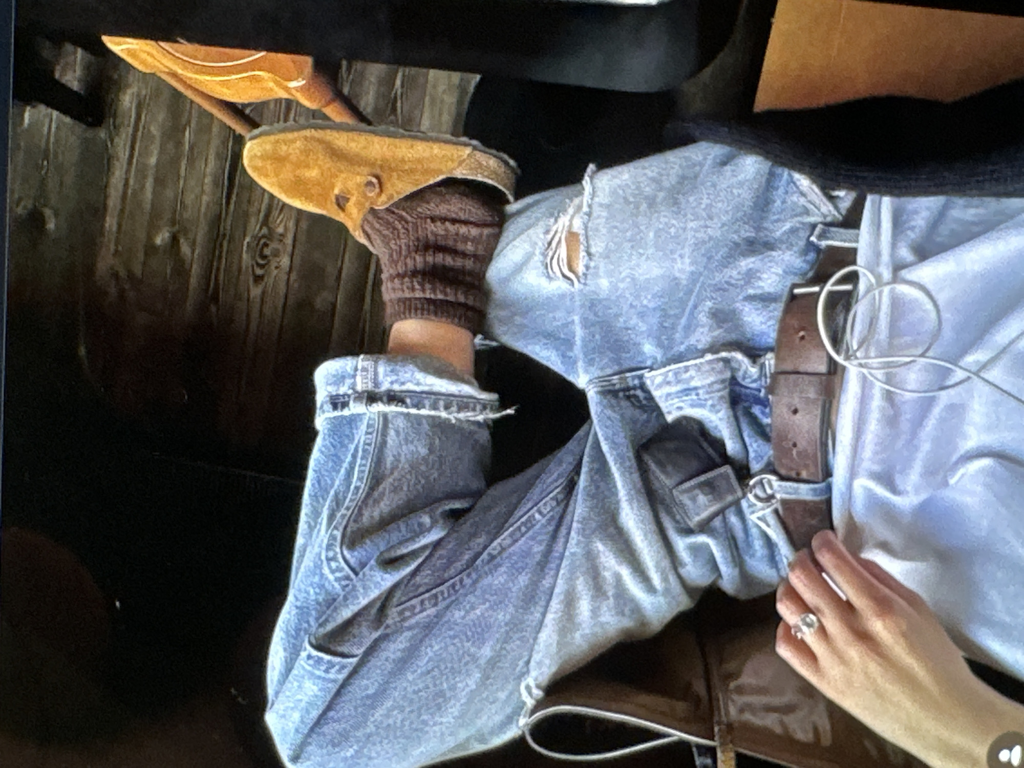 A person wearing light blue, ripped jeans and brown Birkenstock sandals with brown knit socks sits in an indoor setting. A brown leather belt with a silver buckle is visible. A hand with a large ring is positioned near the belt. The location is not discernible from the image. No specific action or interaction is evident.