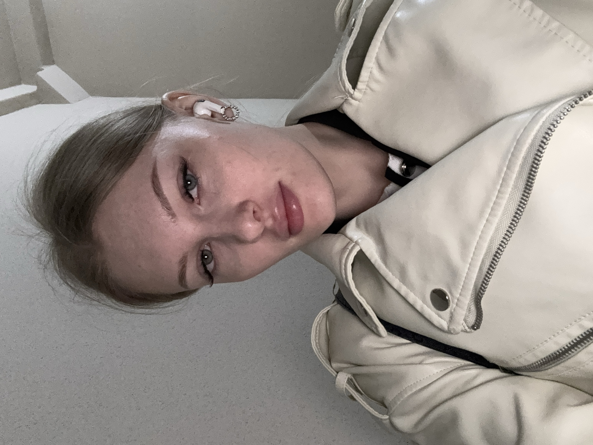 A fair-skinned young woman with light brown hair pulled back is wearing a light-colored jacket and an earbud in her left ear. She is in Perm, Russia. The jacket is zipped and has a visible zipper. The background is a light gray surface. A staircase is visible in the distance.