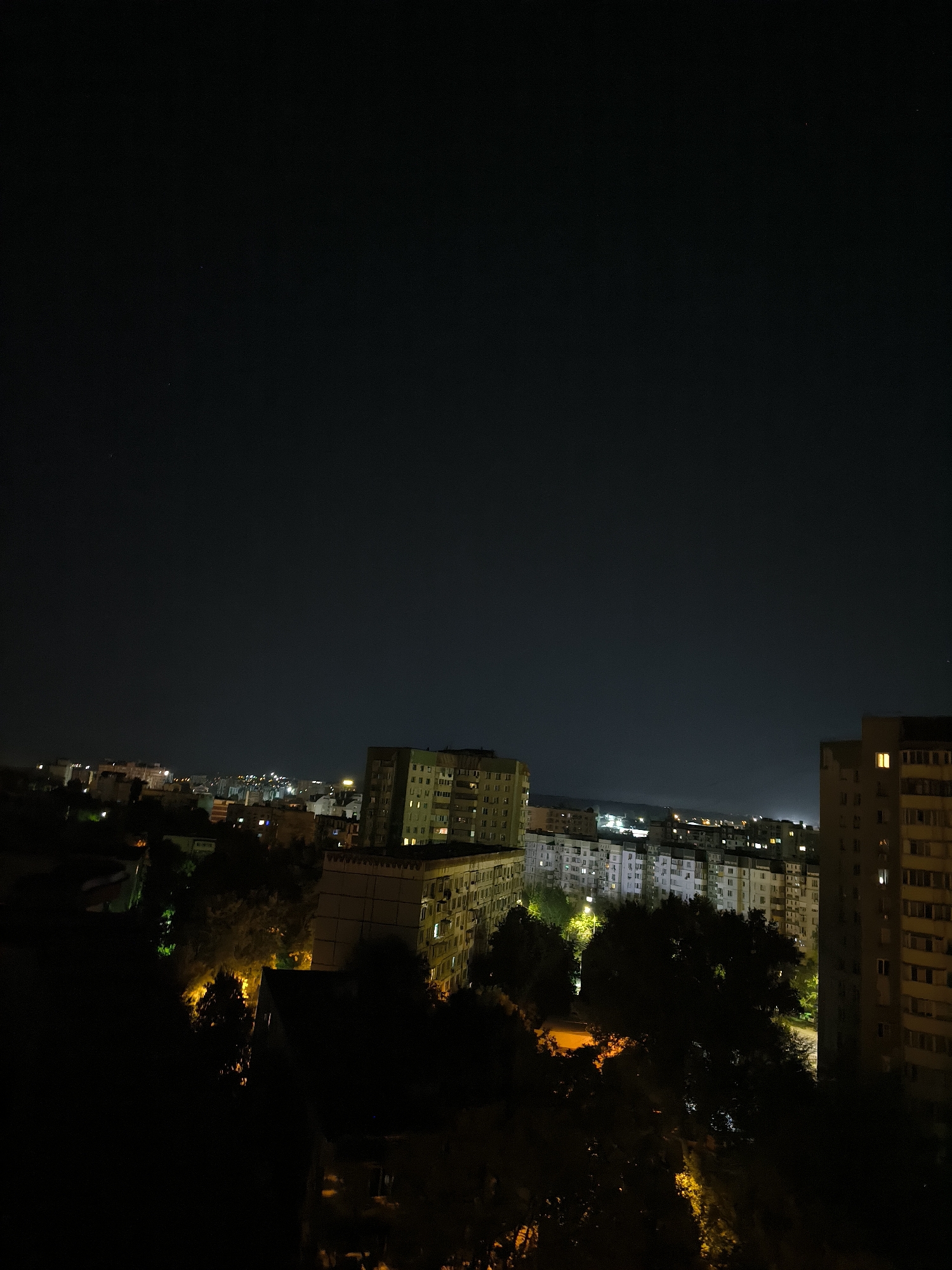 This is a nighttime, wide-angle shot of a cityscape in Chișinău, Moldova. The sky is dark with a few faint stars visible in the upper portion of the frame. The majority of the image is dominated by a dense cluster of apartment buildings, many of which are illuminated from within, suggesting that people are inside.
In the foreground and mid-ground, several multi-story apartment buildings, characteristic of Soviet-era architecture, are prominently featured. These buildings appear to be constructed from concrete and have numerous windows. Some of these windows glow with a warm yellow light, while others are dark. Trees are interspersed among the buildings, their dark foliage contrasting with the illuminated windows. Streetlights and the lights from the buildings cast a warm glow on the surrounding area, particularly in the lower half of the image where the density of trees is higher.
The buildings vary in height and design, creating a layered effect across the urban landscape. The overall impression is of a residential area at night, with the city alive with activity behind the darkened facades of the buildings. There are no people visible in the image. The atmosphere is quiet and still, typical of a city at night.