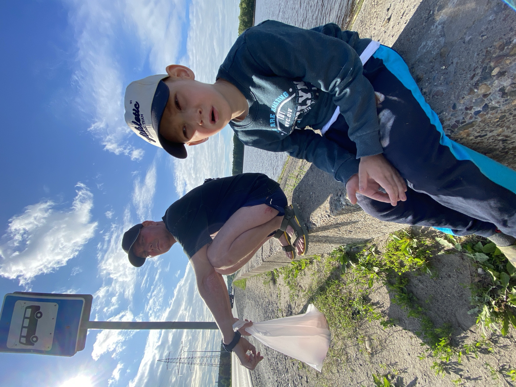 In Noviy, Russia, two individuals, an adult male and a young boy, are seated near a bus stop sign by a body of water. The man, wearing a black t-shirt and shorts, squats with a white plastic bag in his hand. The boy, in a dark sweatshirt and dark pants with blue accents, sits beside him, looking towards the camera. The scene is outdoors with a partly cloudy sky and sunlight. A road is visible behind the man, with power lines and trees in the distance. The ground is a mix of dirt, gravel, and grass, and they are seated on a concrete barrier.