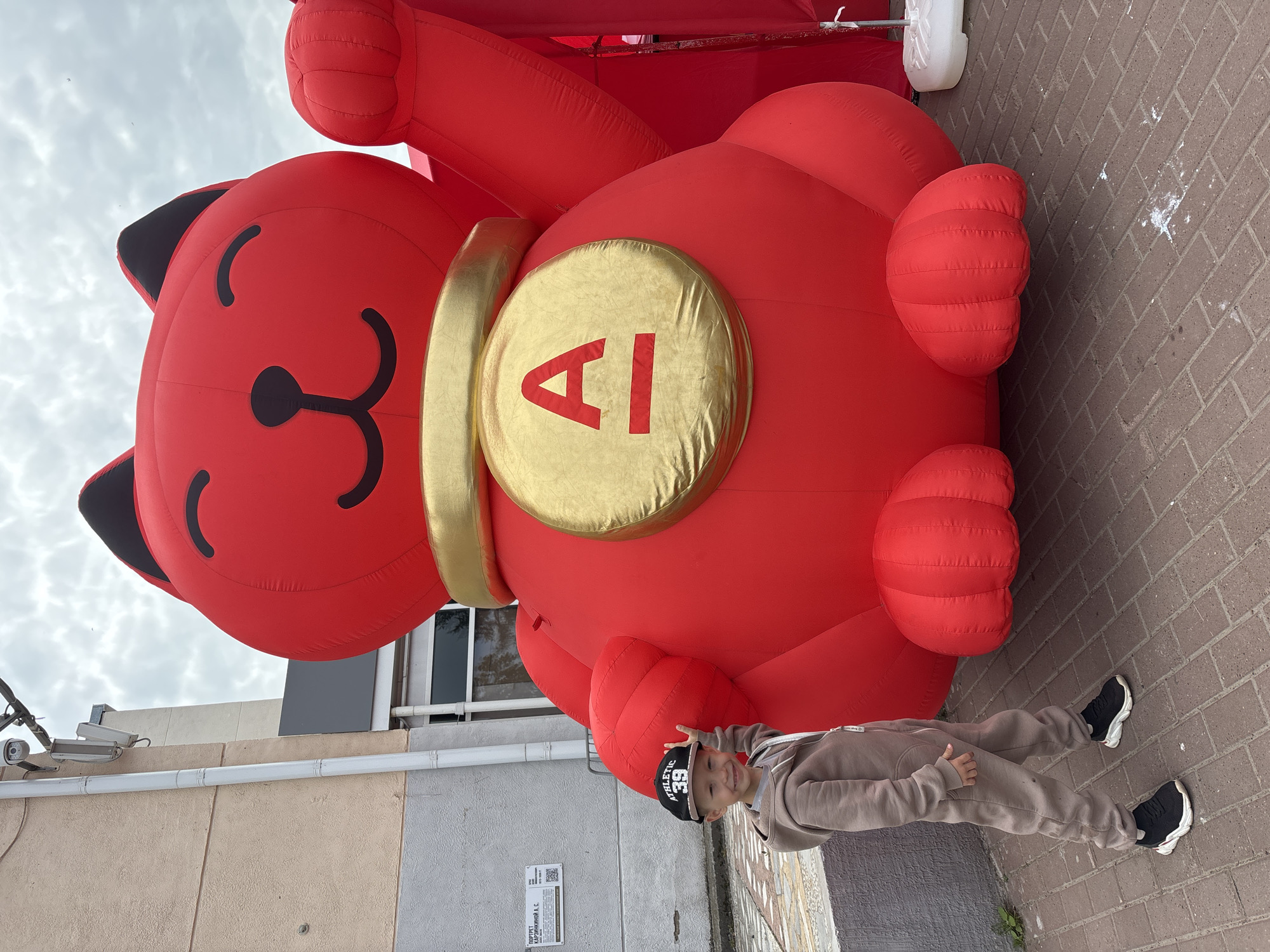 A young boy wearing a beige tracksuit and a black baseball cap stands next to a large, inflatable red cat with a golden collar and a gold circle emblem displaying a red letter "A" on its chest. The boy is smiling and pointing towards the inflatable cat. The cat is positioned outdoors on a brick-paved area. In the background, there is a building with a light-colored facade, windows, and a security camera mounted on a pole. The sky is overcast. The image was taken in Chaikovsky, Russia.