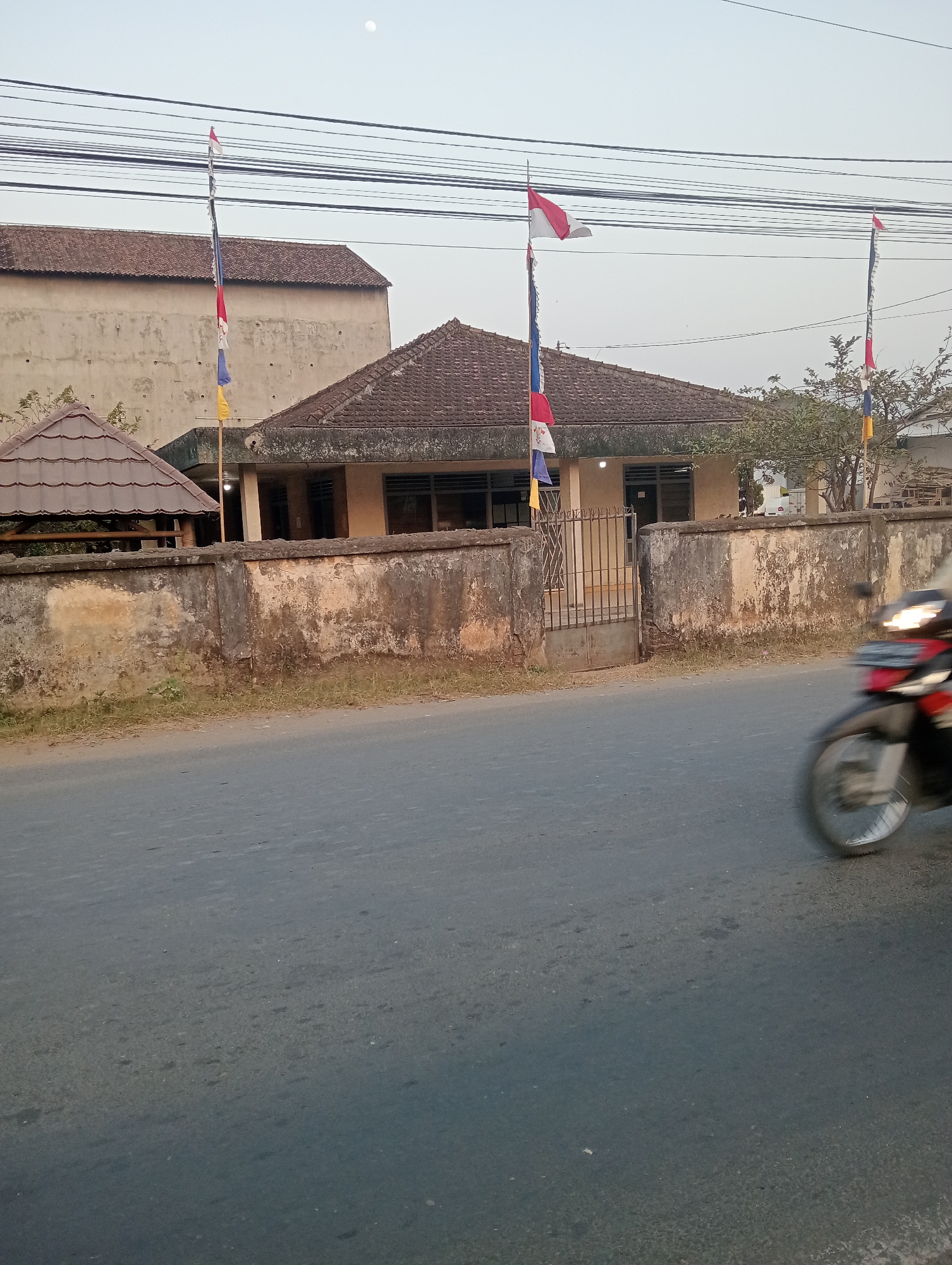 The image depicts a street scene in Pringsewu, Indonesia, with a building behind a weathered wall. Several Indonesian flags are prominently displayed on flagpoles in front of the building. A motorcycle is captured in motion on the right side of the frame, its front wheel blurred due to movement. The building appears to be a single-story structure with a tiled roof and several windows. The wall in front of the building is old and shows signs of wear and tear. The sky is overcast, suggesting it may be dawn or dusk.