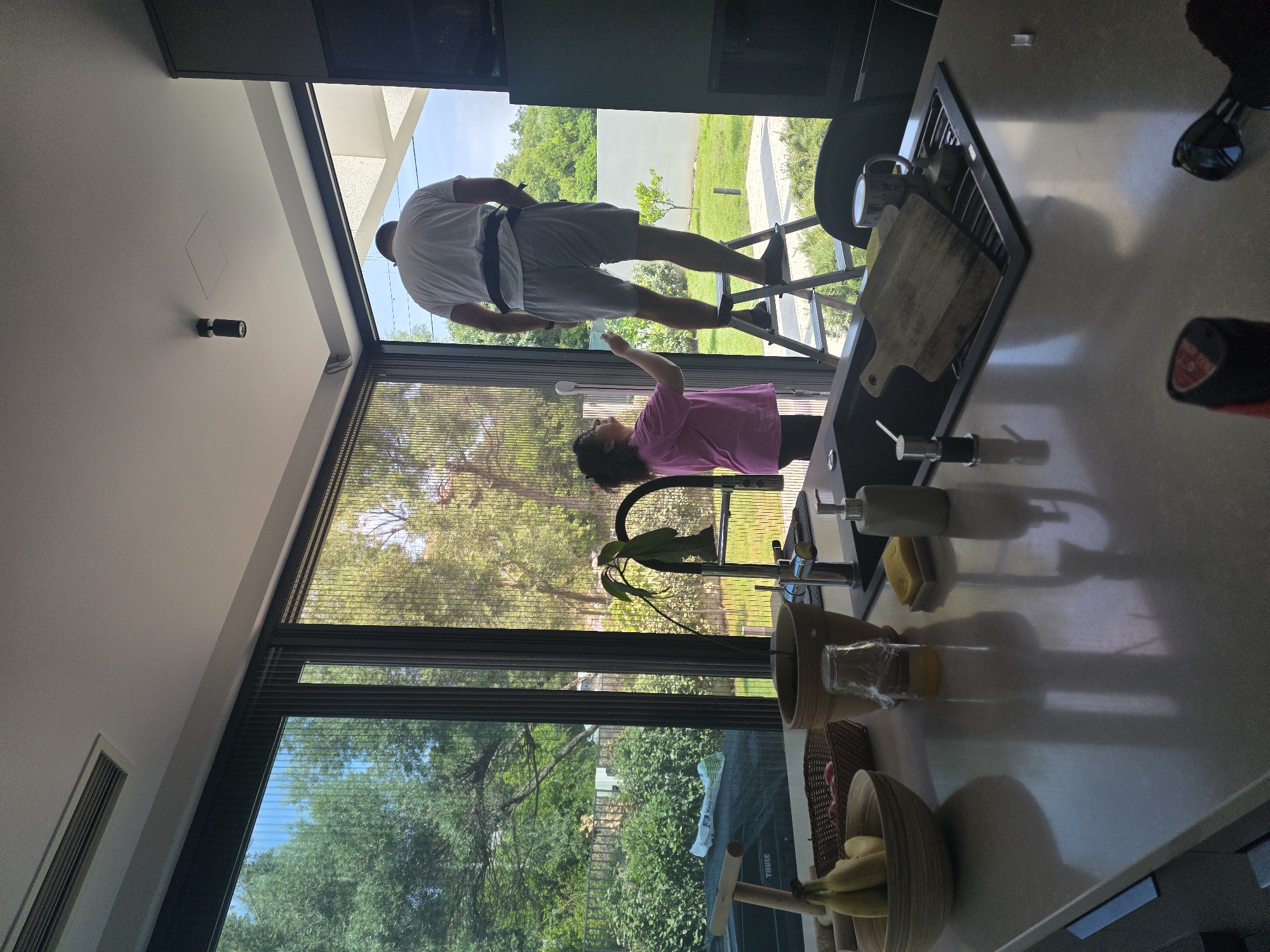 In a modern kitchen in Bogišići, Montenegro, a man wearing a white t-shirt and grey shorts stands on a stepladder, reaching towards the ceiling. He is wearing a black belt-like harness around his waist. Beside him, a woman in a pink t-shirt and black leggings holds a cleaning tool, also appearing to be cleaning the area near the ceiling. Large windows in the background offer a view of lush green trees and a bright sky. The kitchen counter in the foreground is made of a light-colored, reflective material and is adorned with various items, including a bowl of fruit, a potted plant, a soap dispenser, a sponge, and a cutting board in the sink. The overall atmosphere suggests a domestic chore or maintenance activity taking place in a well-lit, contemporary living space.