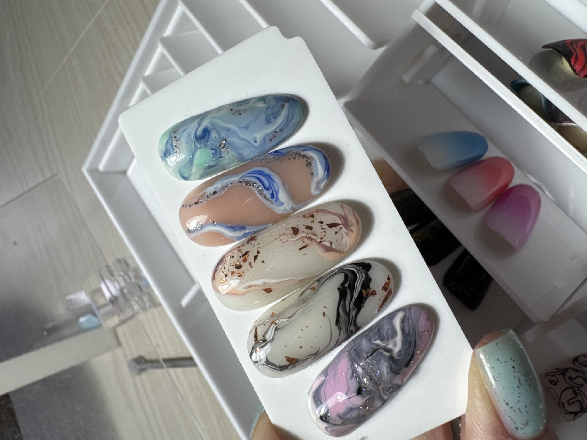 The image shows a close-up overhead view of several artificial nails with marble-like designs. The nails are arranged in a white tray, with five prominent nails on display in the foreground. These nails feature swirling patterns in various color combinations, including pink and gray, black and white with copper flakes, beige with copper flakes, peach with blue and white swirls, and blue and green with silver glitter. A hand is holding the lower left portion of the tray, revealing a fingernail painted in a pale green with subtle glitter. In the background, partially visible within the white tray, are more artificial nails with gradient color designs, transitioning from pink to blue, and other nails with intricate patterns. The lighting suggests an indoor setting, possibly a beauty salon or a personal nail art station. The background also contains a partially visible clear bottle with a silver cap, likely a nail polish or top coat. The overall scene focuses on the artistry and detail of the nail designs.