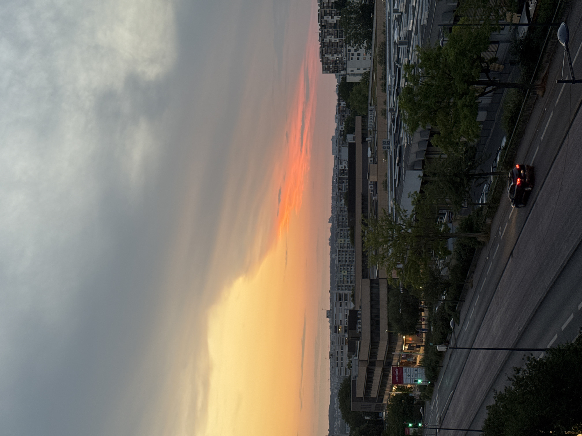 This is a landscape, aerial, medium shot of a city street at dusk, with a sunset in the background. The sky is a mix of gray clouds and vibrant orange and pink hues. In the foreground, a multi-lane road runs horizontally across the frame, with a few cars driving on it. There are streetlights and traffic lights visible along the road. The middle ground features several buildings, including a large, multi-story office building with a sign that reads "cerisole". There are also trees lining the street and surrounding the buildings. In the background, a dense urban skyline with apartment buildings and other structures stretches to the horizon. The overall scene suggests an urban environment during a sunset.