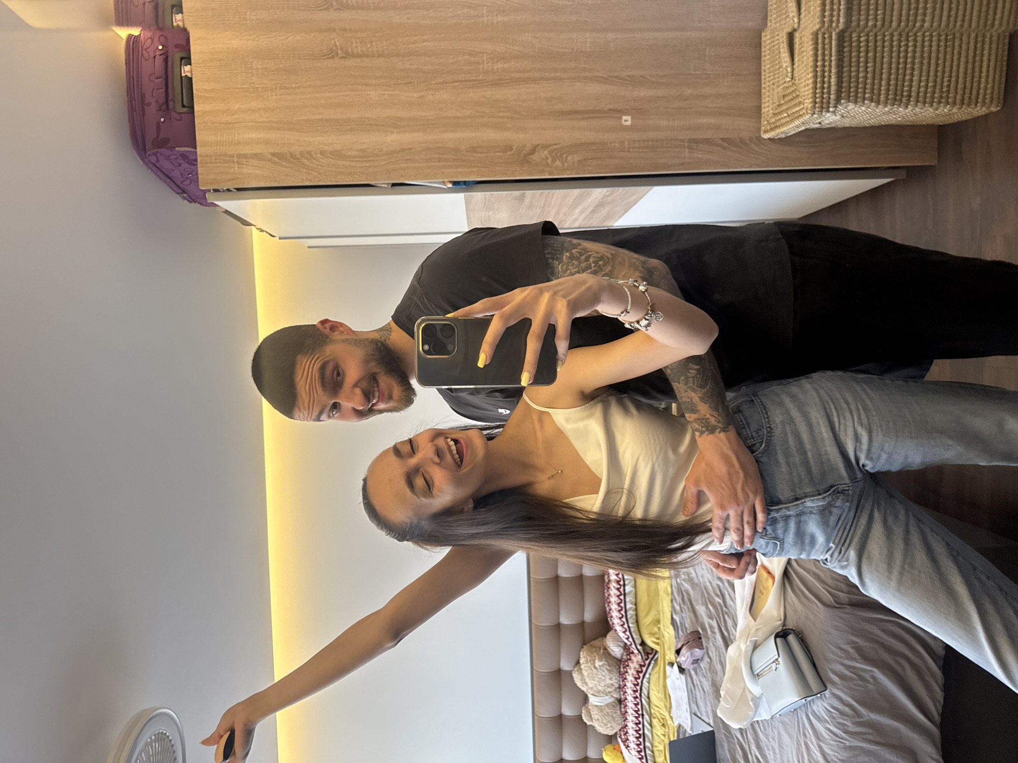 A man and woman are taking a selfie in a room. The woman is holding a phone with yellow accents, and the man has his arm around her. The room contains a bed with a tan headboard, bedding, and a light blue handbag on it. A suitcase sits atop a wooden cabinet. The location appears to be a hotel room in Sofia, Bulgaria, given the style of the furniture and decor.
