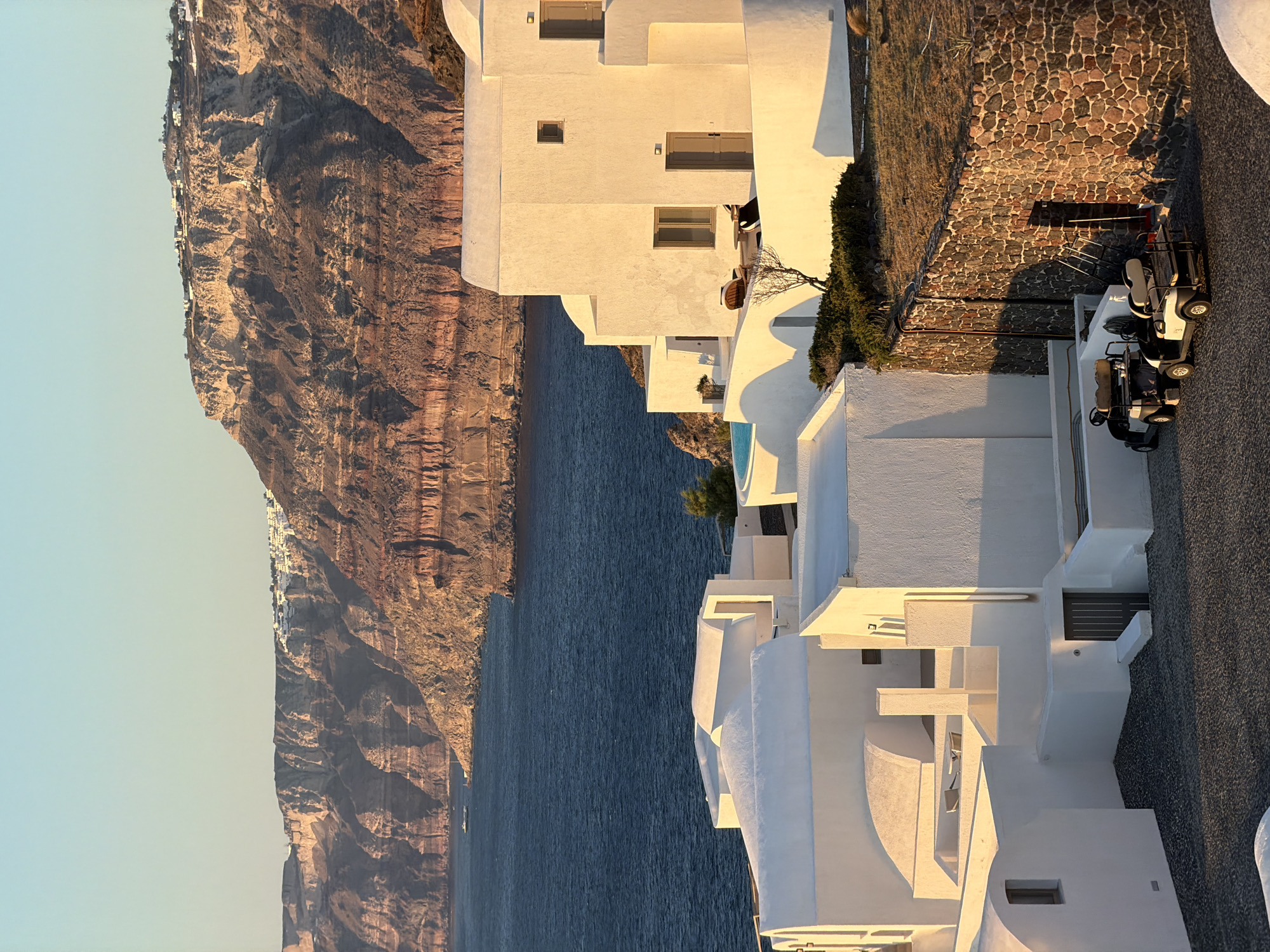 The image shows a view of Oia, Santorini, Greece, with the iconic white-washed architecture cascading down the cliffside towards the Aegean Sea. The foreground features several white, cubic buildings with rounded edges, characteristic of Cycladic architecture. Some of these buildings have small windows and doors, and a few are adorned with decorative elements like arches and sloped roofs. In the middle ground, a stone wall with varied colored rocks encloses an area with dry grass. Two golf carts are parked on a gravel surface in front of the wall. The background reveals the deep blue sea meeting a rugged, layered cliff face, topped with more white structures and buildings that form the village of Oia. The sky is clear and a pale blue, with the warm glow of sunset illuminating the scene.