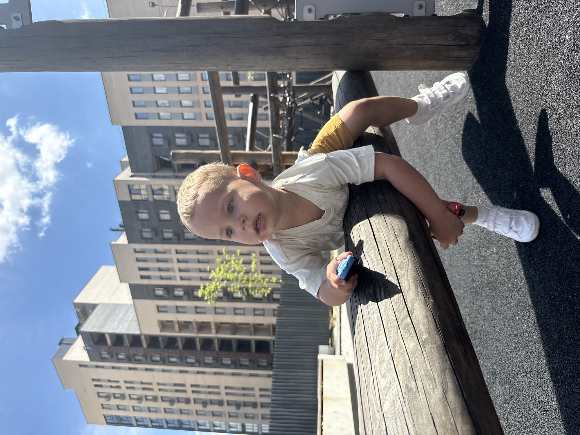 In an outdoor playground setting with modern apartment buildings in the background, a young Caucasian boy with blonde hair is positioned on a wooden play structure. He is wearing a white t-shirt and mustard-colored shorts, along with white sneakers. The boy is holding a small blue toy car in his right hand and appears to be looking to his left, with his body leaning forward on the wooden beam. The ground beneath the play structure is a dark, textured surface, likely rubberized safety surfacing. The scene is lit by bright sunlight, casting distinct shadows.