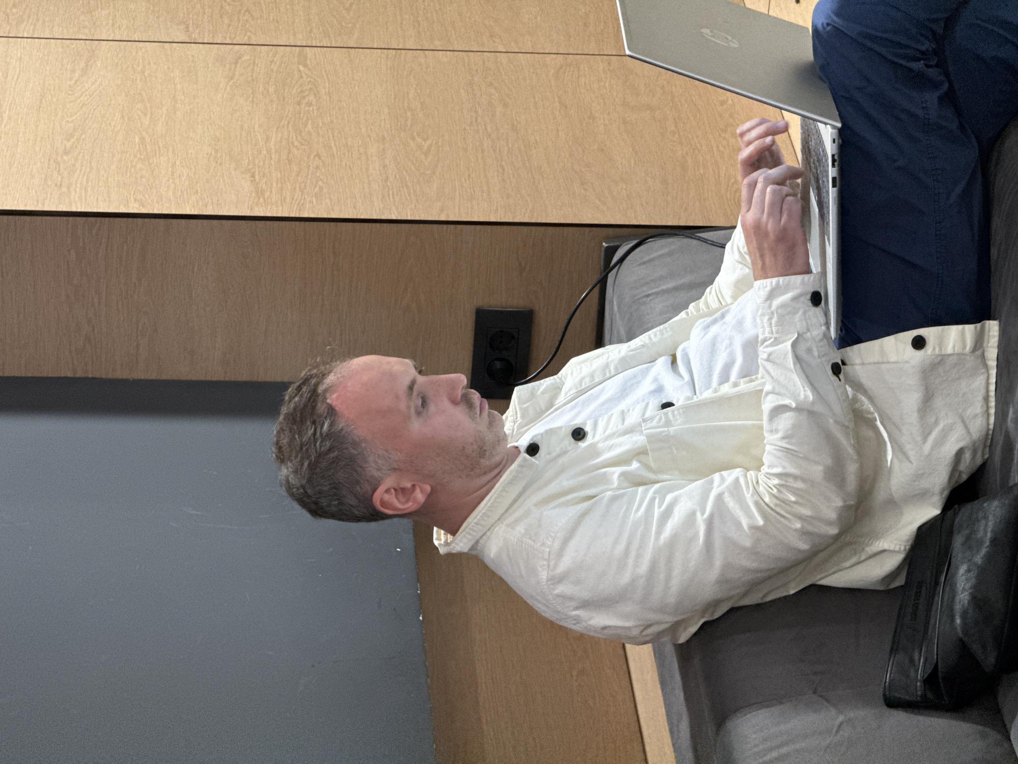 A man is sitting on a gray couch, using a silver laptop. He is wearing blue pants and a white shirt and jacket. Next to him is a black object, possibly a bag. The background includes a wooden panel and a gray wall with a black electrical outlet. The location is Surcin Urban Municipality, Serbia.