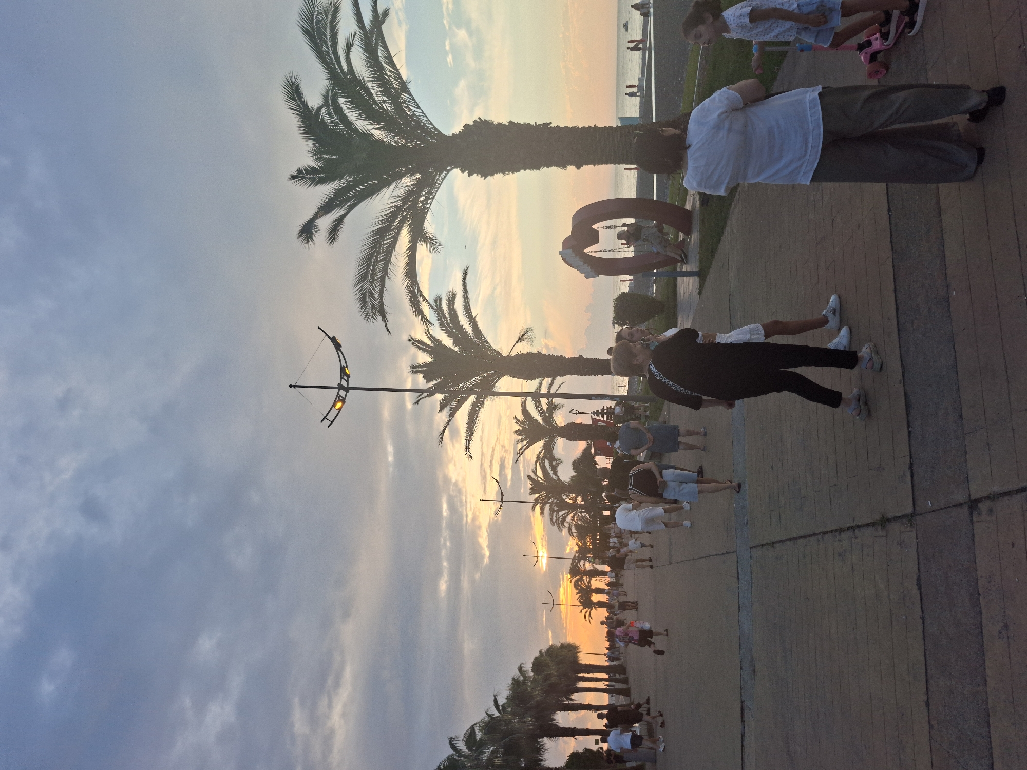 The image depicts a sunset scene along Batumi's Boulevard in Georgia. A paved walkway, lined with palm trees, is filled with pedestrians of various ages enjoying the evening. In the foreground, several individuals are walking or standing, with a woman in a white shirt and olive pants facing away from the camera, and a young girl on a pink scooter to her right. Further down the walkway, a group of people are strolling, and in the middle ground, a large heart-shaped sculpture with a swing is visible. The sky is filled with dramatic clouds, tinged with the warm hues of the setting sun. The Black Sea is visible in the distance, with a few people on the pebble beach. Streetlights illuminate the pathway, indicating the transition from day to evening.