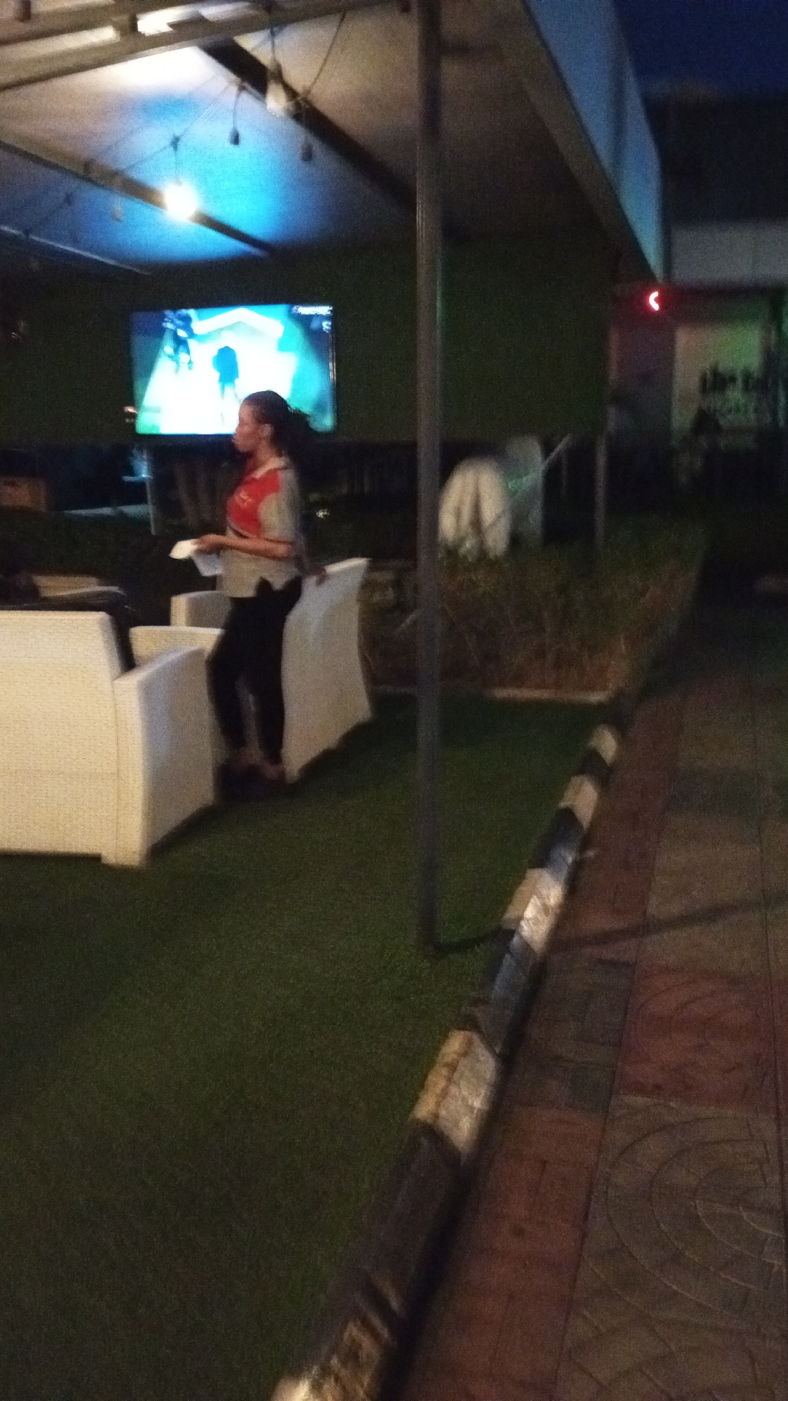 The image depicts an outdoor seating area in what appears to be a restaurant or bar. A woman in a red and gray shirt and black pants stands near white cushioned chairs. A large television screen displaying brightly colored graphics is visible behind her. A fence with small bushes is partially visible behind the chairs, and a black and white striped border delineates the edge of a walkway. The flooring is artificial grass. A metal support structure is in the foreground. The lighting is dim, with some overhead lights. Based on the prompt, the location is Adankolo, Nigeria.