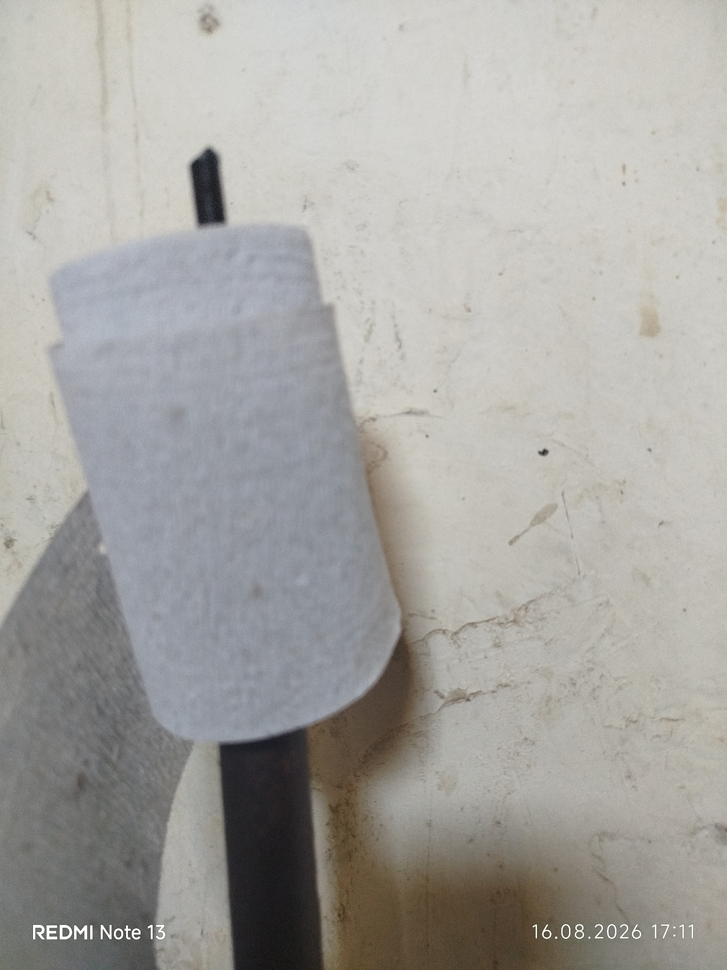 The image shows a close-up view of a textured, off-white wall with some staining and discoloration. A black, cylindrical object protrudes from the top of the frame, and a light gray, cylindrical object with a textured surface is partially visible, surrounding the black object. The bottom left corner of the image displays the text "REDMI Note 13", and the bottom right corner shows the date and time "16.08.2026 17:11". The scene does not provide any specific visual cues to indicate its location in Kungirat, Uzbekistan.