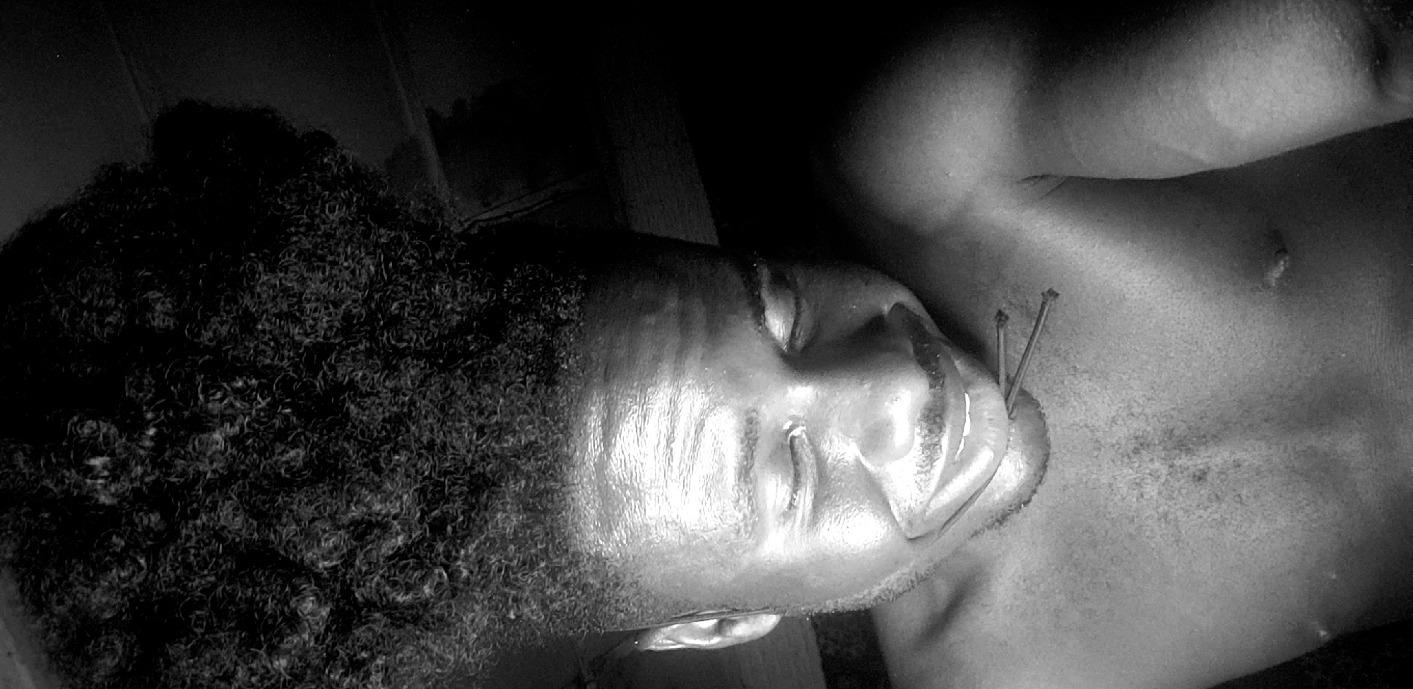 A black and white image depicts a person lying down, their head resting on what appears to be a hard surface. The person has dark, curly hair and appears to be asleep. Two thin, dark objects, possibly nails or needles, are protruding from the right side of their cheek. The image is dimly lit, and the surrounding environment is unclear. Location provided is Narayi, Nigeria.