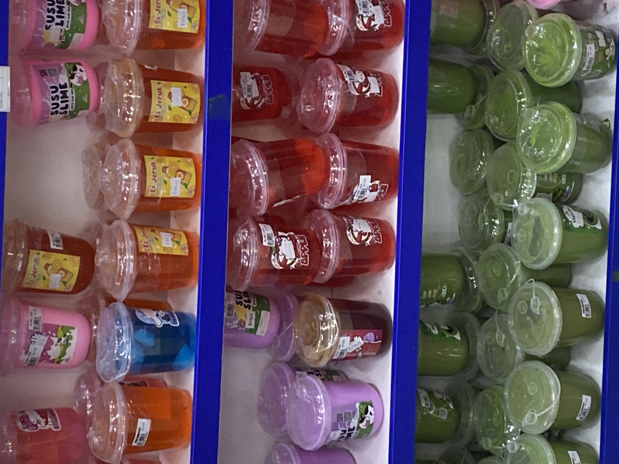 The image displays a retail setting, likely a shelf in a store in Depok, Indonesia. The shelves are stocked with various plastic containers filled with brightly colored liquids and slimes. The top shelf shows pink containers labeled "SUSU SLIME" featuring cartoon cows, and orange containers with "Es Jeruk" labels depicting sliced oranges. Below, there are translucent orange and blue-filled cups, also with "Es Jeruk" labels. Further down, purple containers with similar branding are visible, alongside red containers labeled "APPLU" with cartoon apple graphics. The bottom shelf is filled with numerous green containers, some with lids and spouts, suggesting they might be beverages or smoothies, with some labeled "Matcha". The overall scene is one of organized product display in a commercial environment.